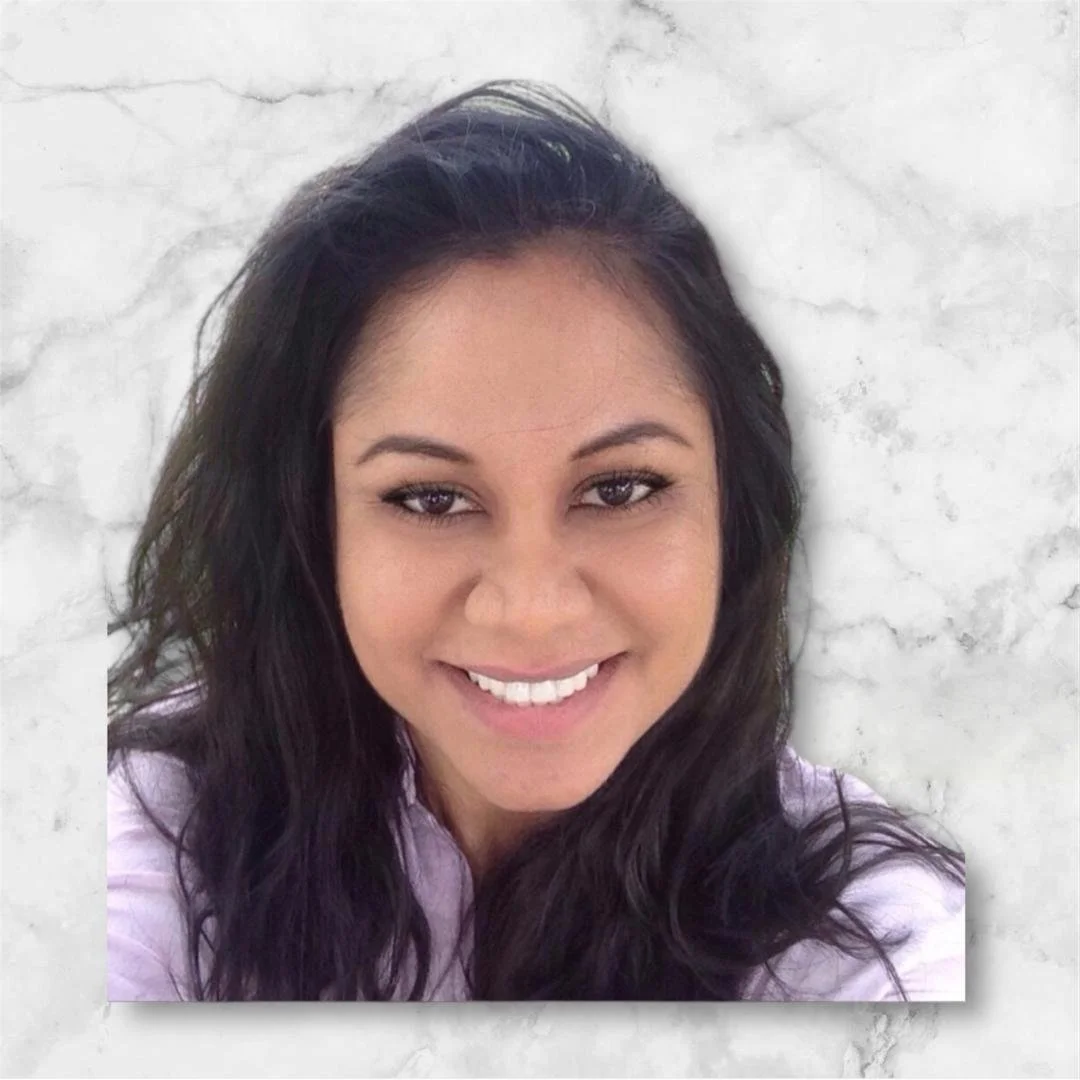 A woman with dark brown hair and light makeup smiling, wearing a light-colored top, against a light stone wall background.