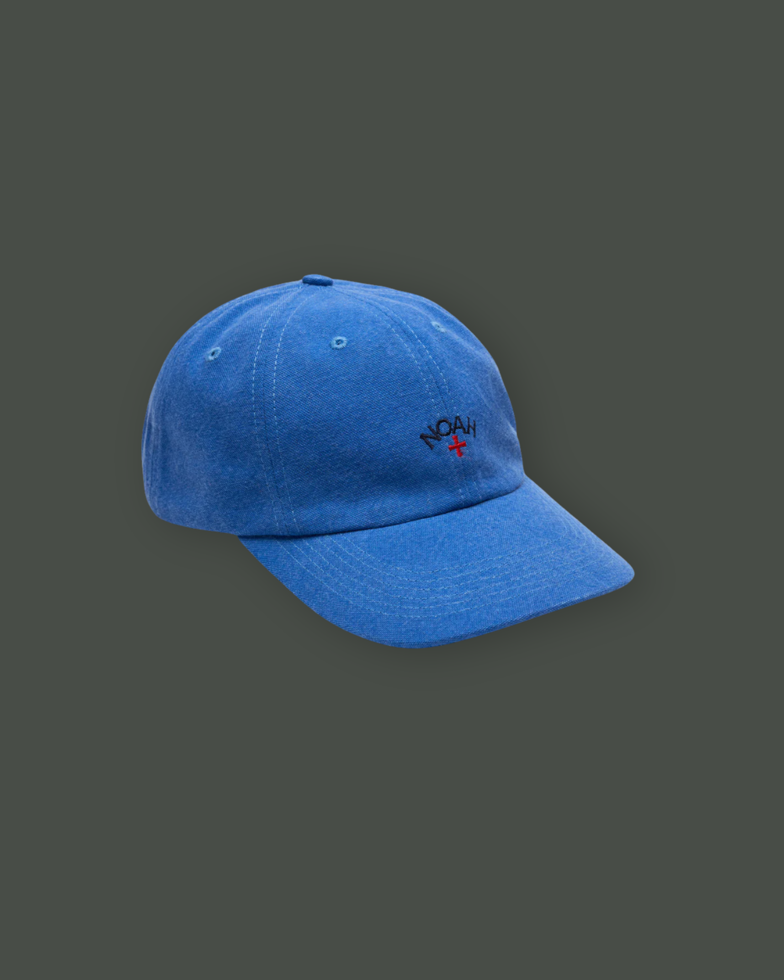 Blue baseball cap with embroidered text 'NOAH' and a small red cross below it, shown against a dark background.