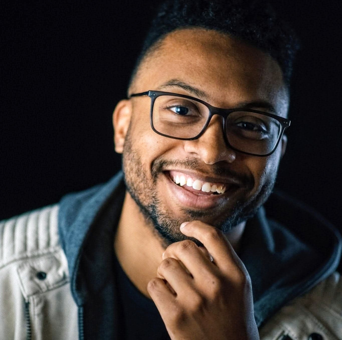 Smiling young man with glasses, beard, and mustache, wearing a dark shirt and light-colored jacket, posing against a black background.