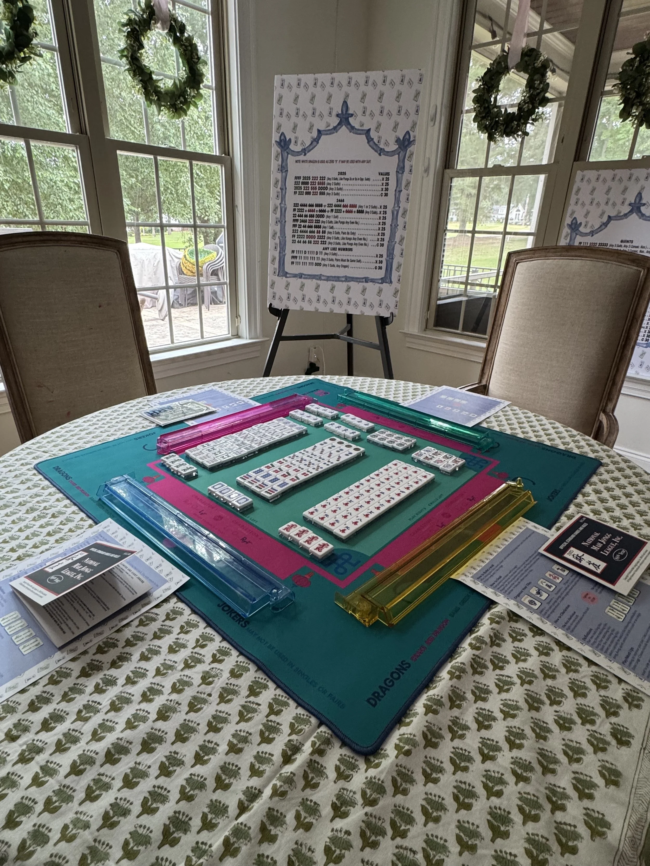 An indoor room set up for playing Mahjong, with a Mahjong board and tiles on a table covered with a patterned tablecloth, and a decorative poster and instructions on easels in the background near windows decorated with wreaths.