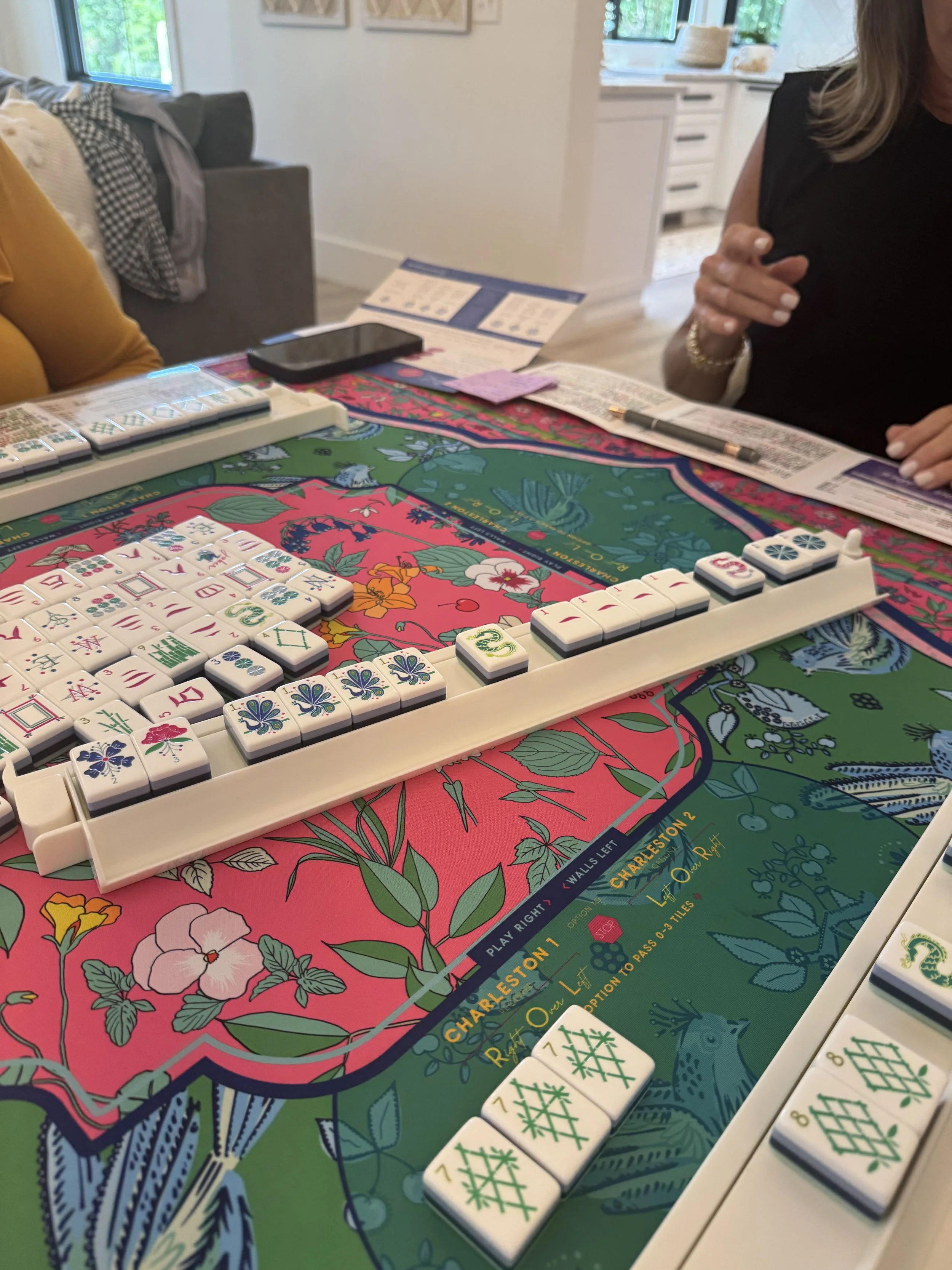 Close-up of a colorful Mahjong game set up on a vibrant floral patterned table. The game tiles are arranged in a row, with some tiles face-up and others face-down. Two people are sitting at the table, one on each side, engaged in the game.