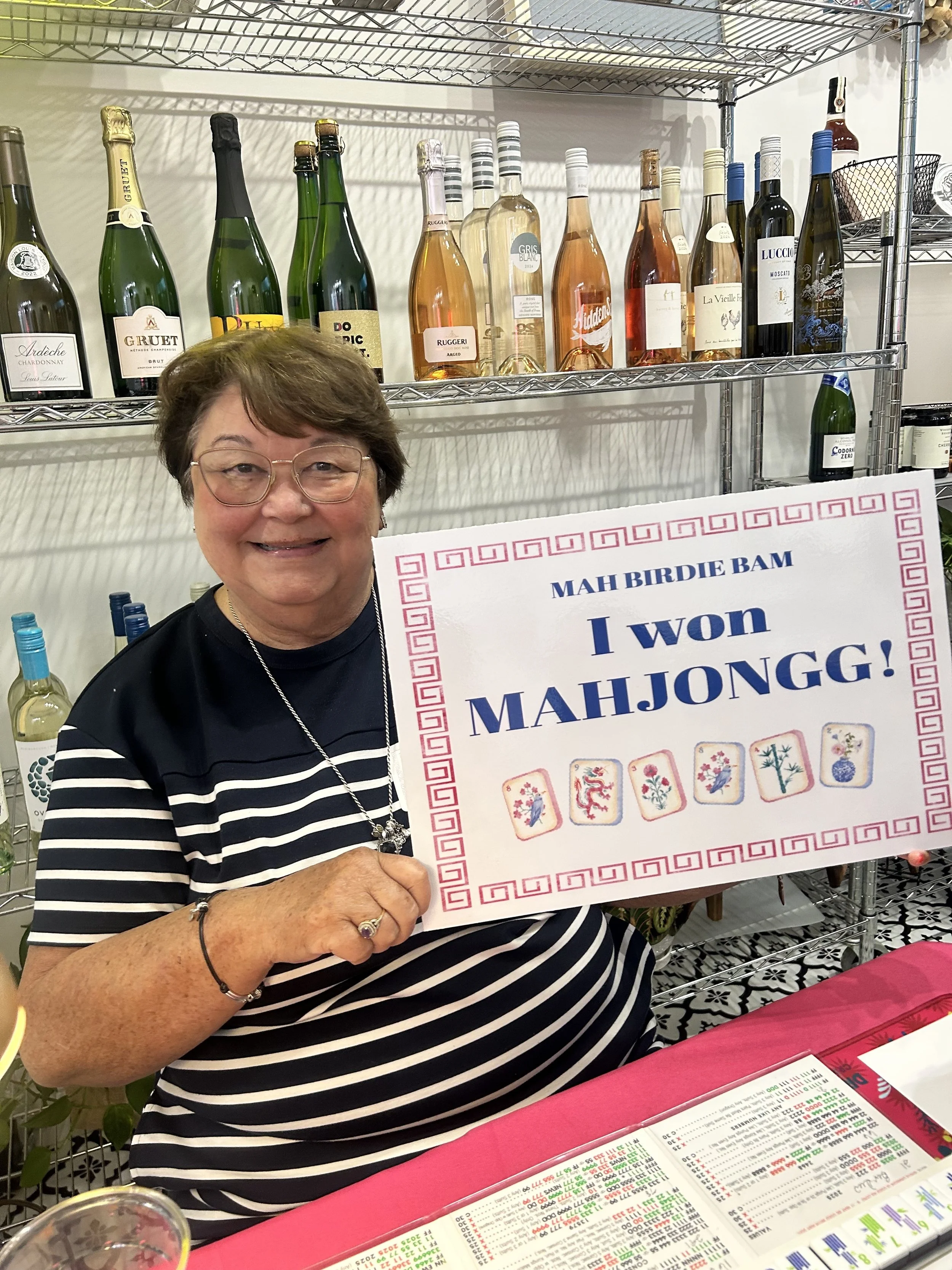 An older woman with glasses and a striped black and white shirt sitting at a table, smiling and holding a sign that says "MAH BIRDIE BAM I won MAHJONGG!" with decorative tiles at the bottom, in a room with shelves of wine bottles in the background.