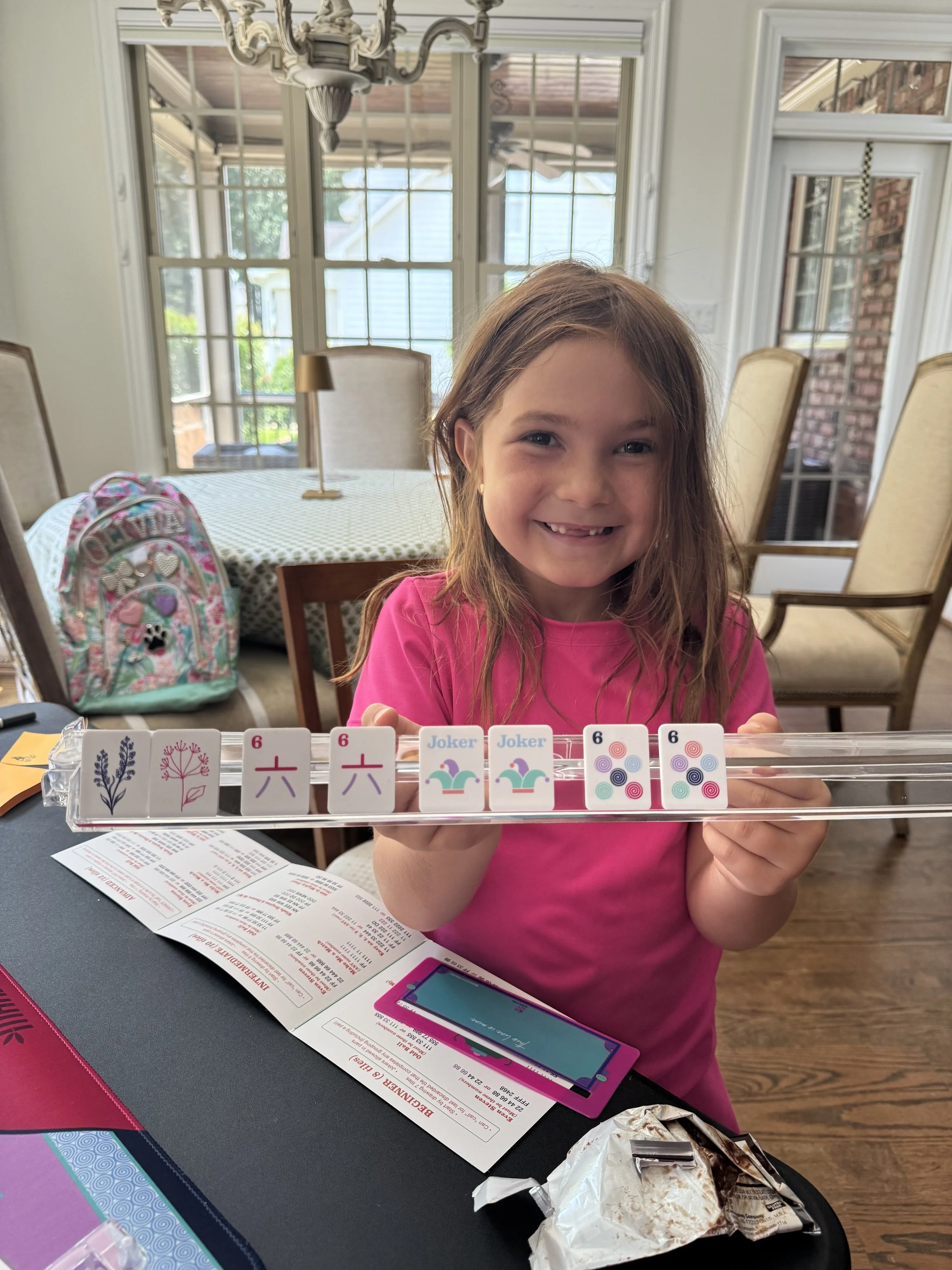 A young girl with long light brown hair wearing a pink shirt, smiling and showing a tile of dominoes she is holding, which has flowers, spirals, and jokers on them, in a bright dining room with large windows.