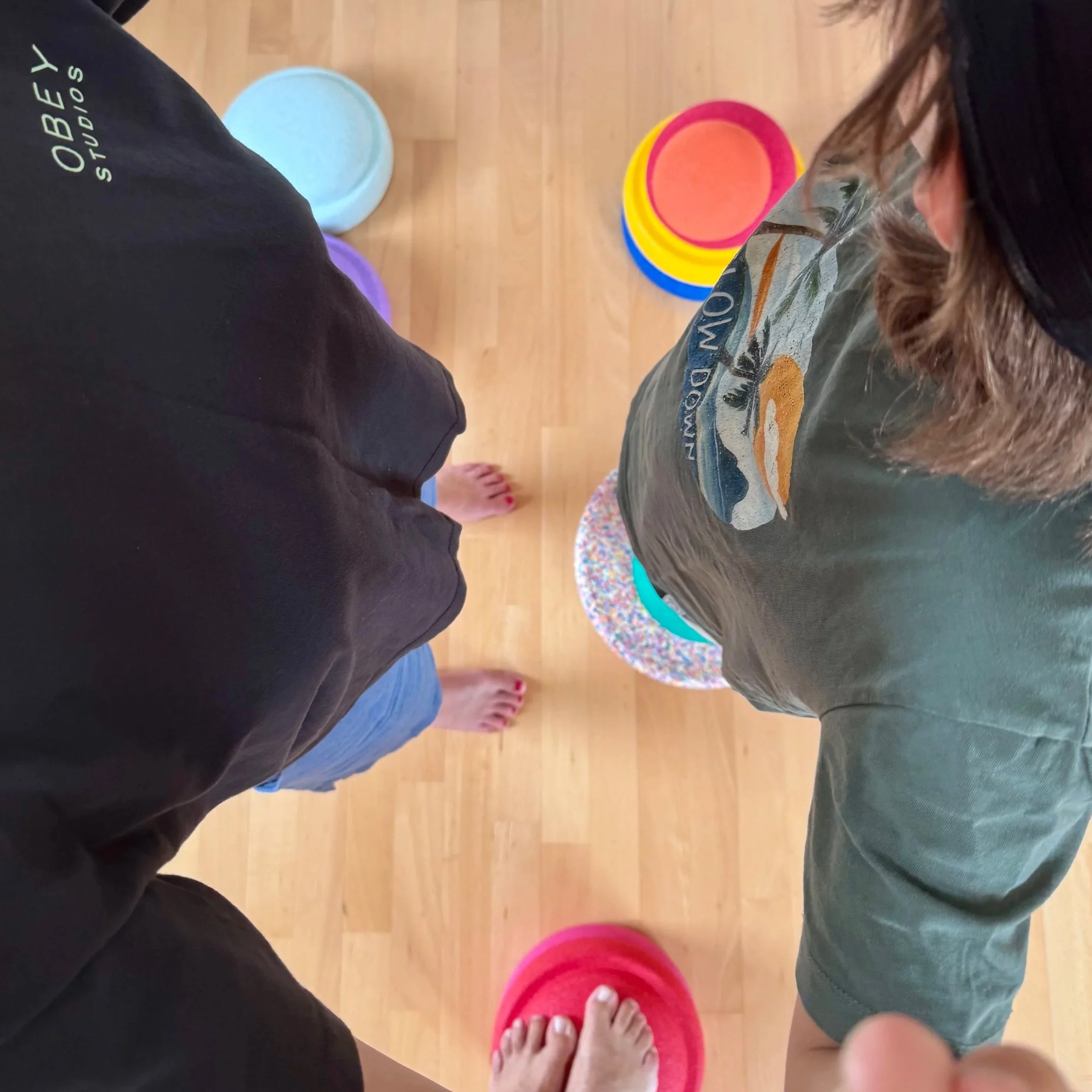Das Foto zeigt mehrere bunte Stapelsteine, die auf einem Holzboden liegen. Zwei Personen stehen auf unterschiedlichen Platten, machen Eltern-Kind-Yoga, nackte Füße sind sichtbar. Die Perspektive ist von oben aufgenommen.