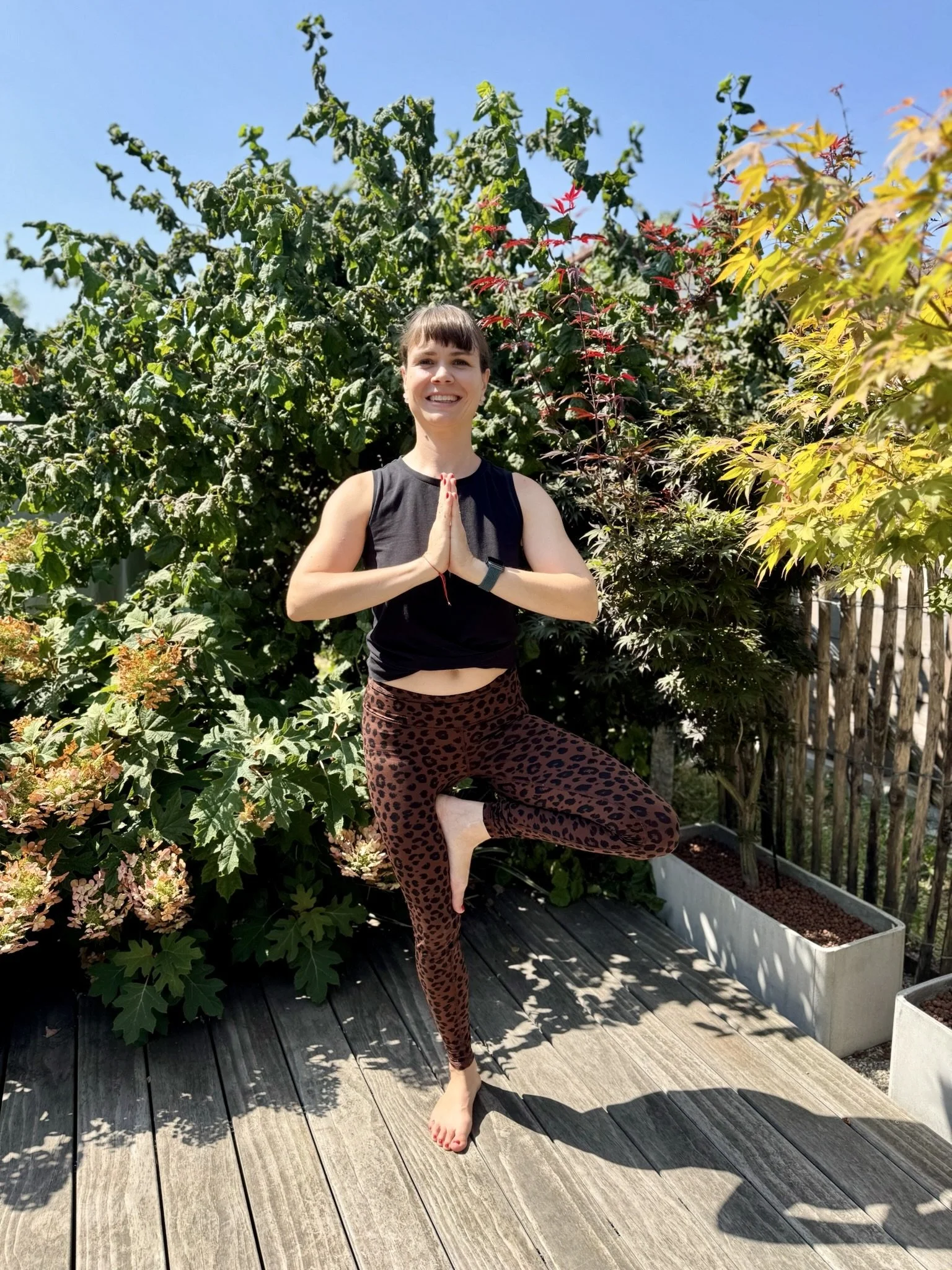 Eine junge Frau beim Yoga im Garten, steht auf einem Bein in der Baumhaltung, umgeben von grünen Büschen und bunten Blättern bei sonnigem Wetter.