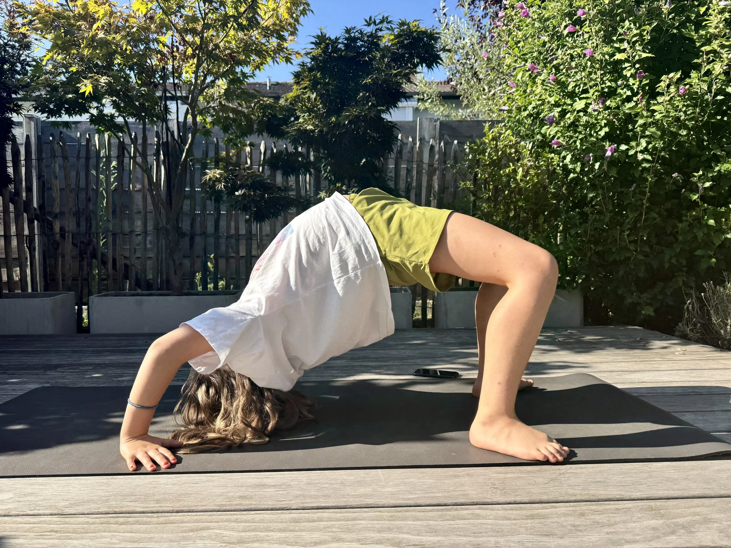 Ein Mädchen macht das große Rad, eine Yoga-Haltung, auf einer Matte im Garten bei Sonnenlicht, umgeben von Bäumen und blühenden Sträuchern.