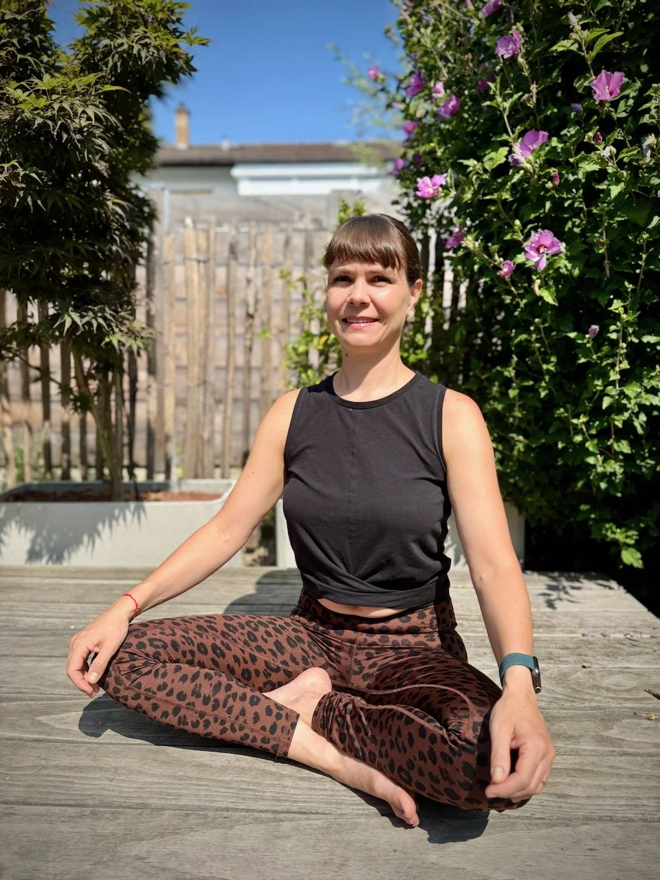 Stefanie Drobnik in der Yoga-Pose Lotussitz im Garten, umgeben von grünen Pflanzen und Blumen, bei Sonnenlicht.