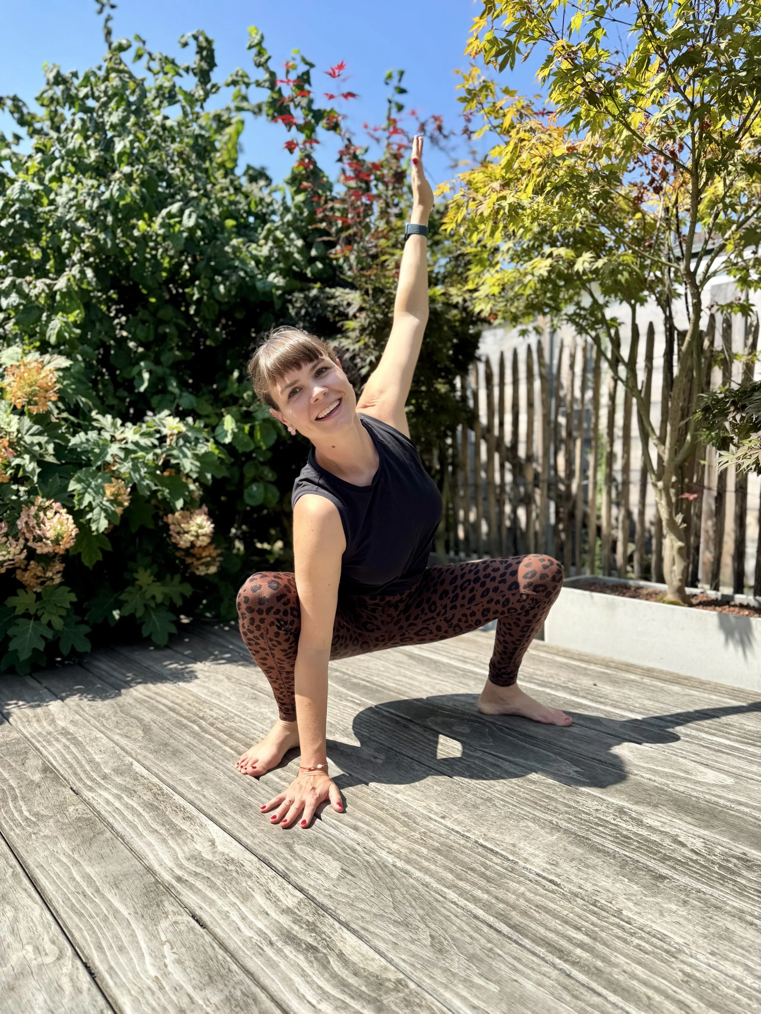 Yogalehrerin macht eine Asana im Freien auf einer Holzterrasse, umgeben von grünen Büschen und Bäumen bei sonnigem Wetter.