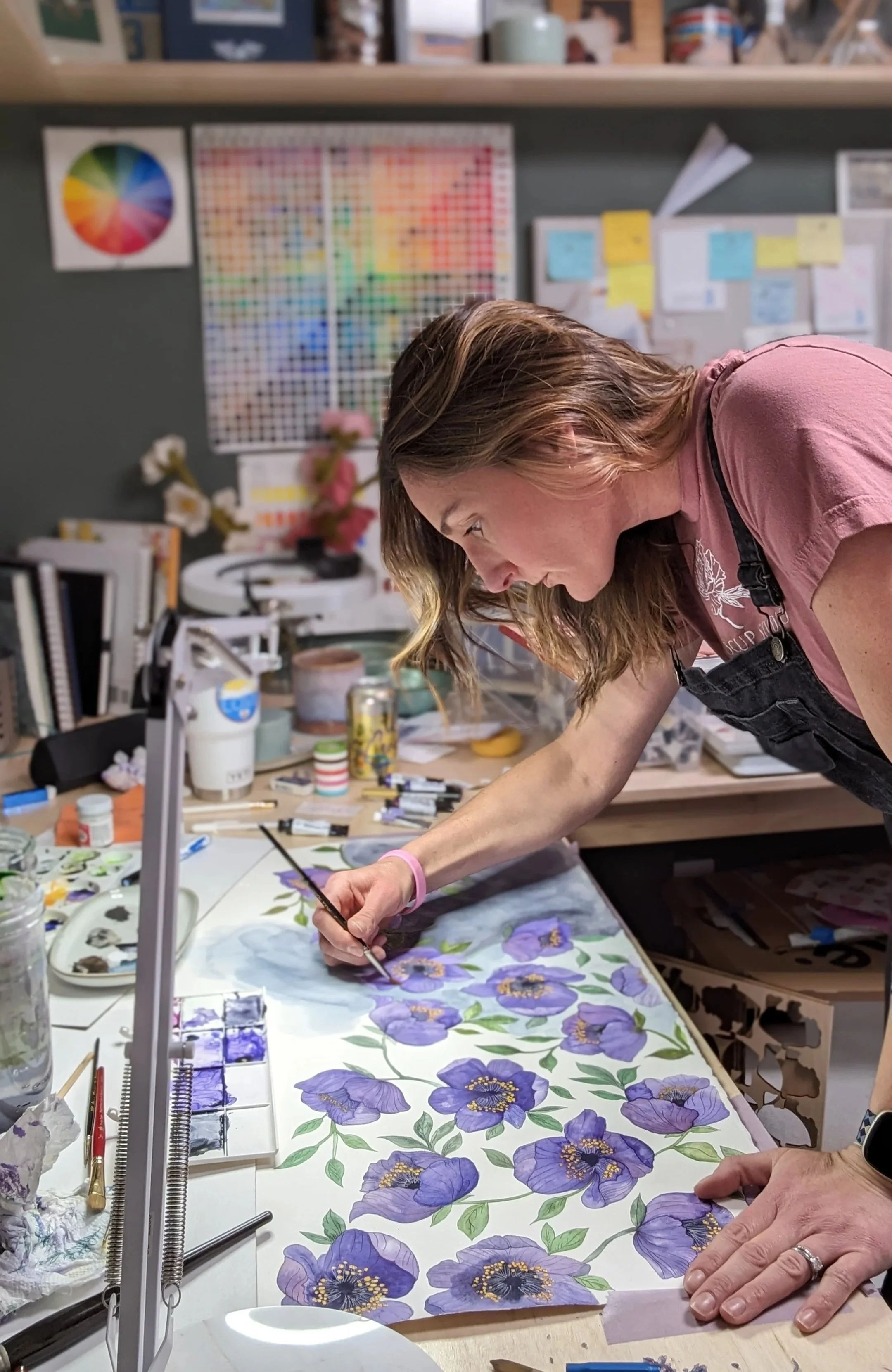 A woman is painting a floral design with purple and yellow flowers on a large sheet of paper in an art studio filled with art supplies and color charts.