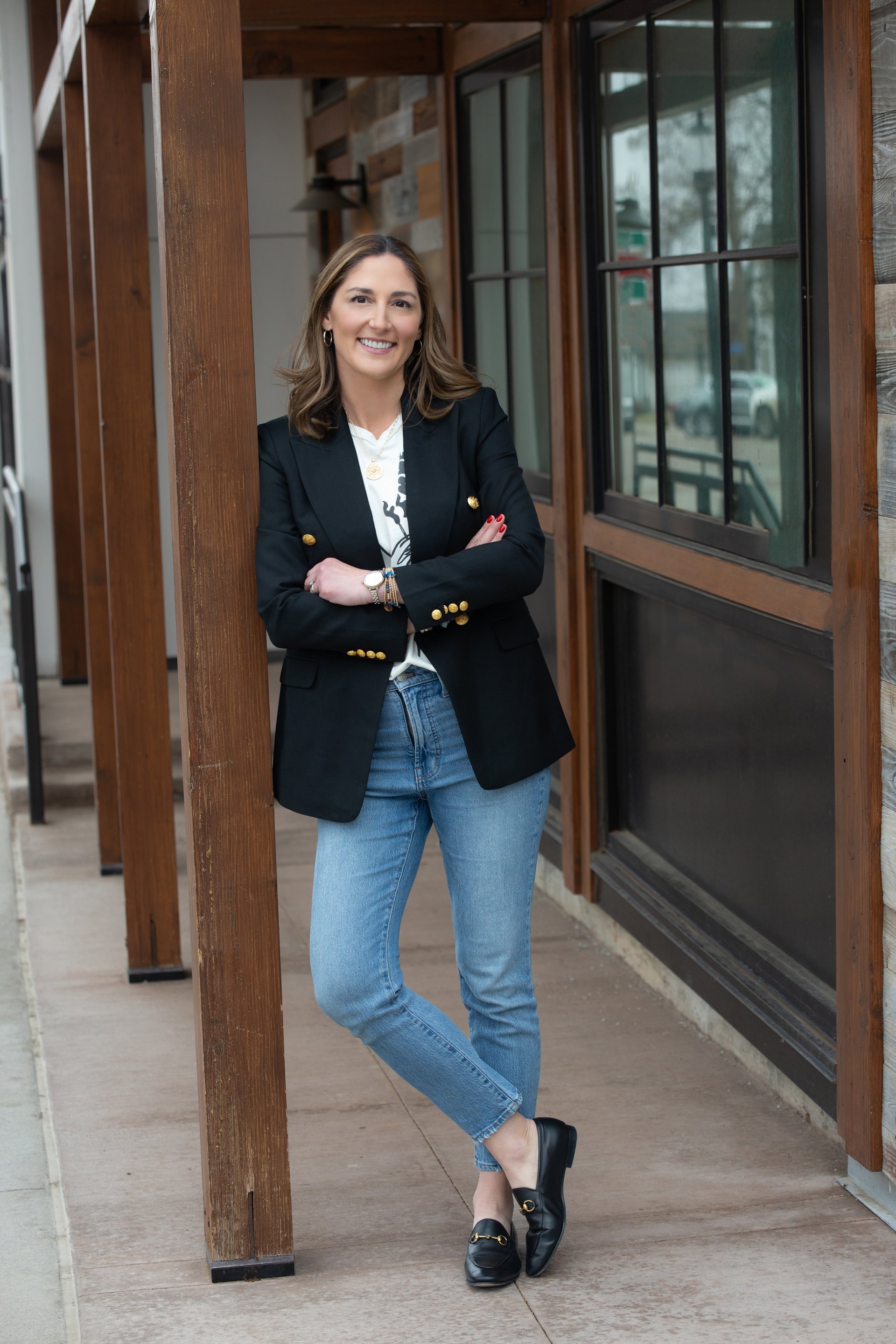 A woman standing outside next to a wooden and glass building, smiling and wearing a black blazer, white shirt, blue jeans, and black shoes.