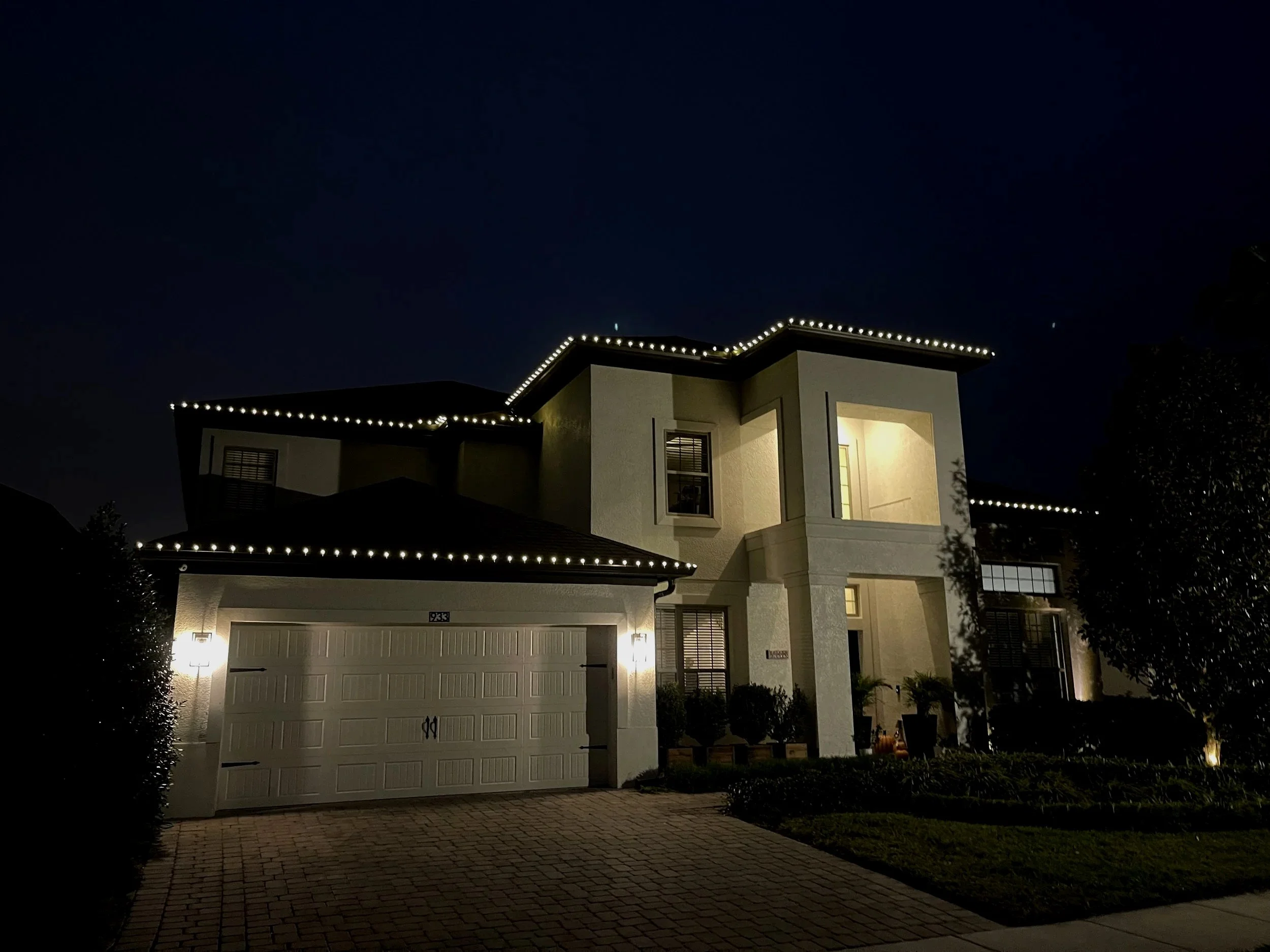 A two-story white house at night with exterior lights and string lights outlining the roof, surrounded by a paved driveway, bushes, and trees.