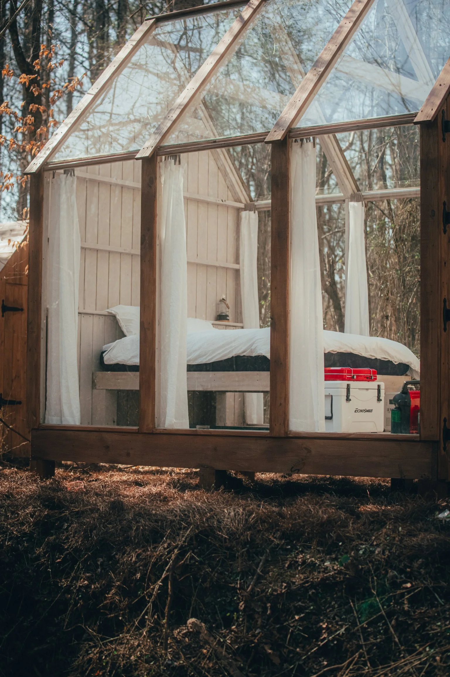 A small wooden and glass house with curtains, housing a bed and outdoor wood fired hot tub, situated in a forrest with trees