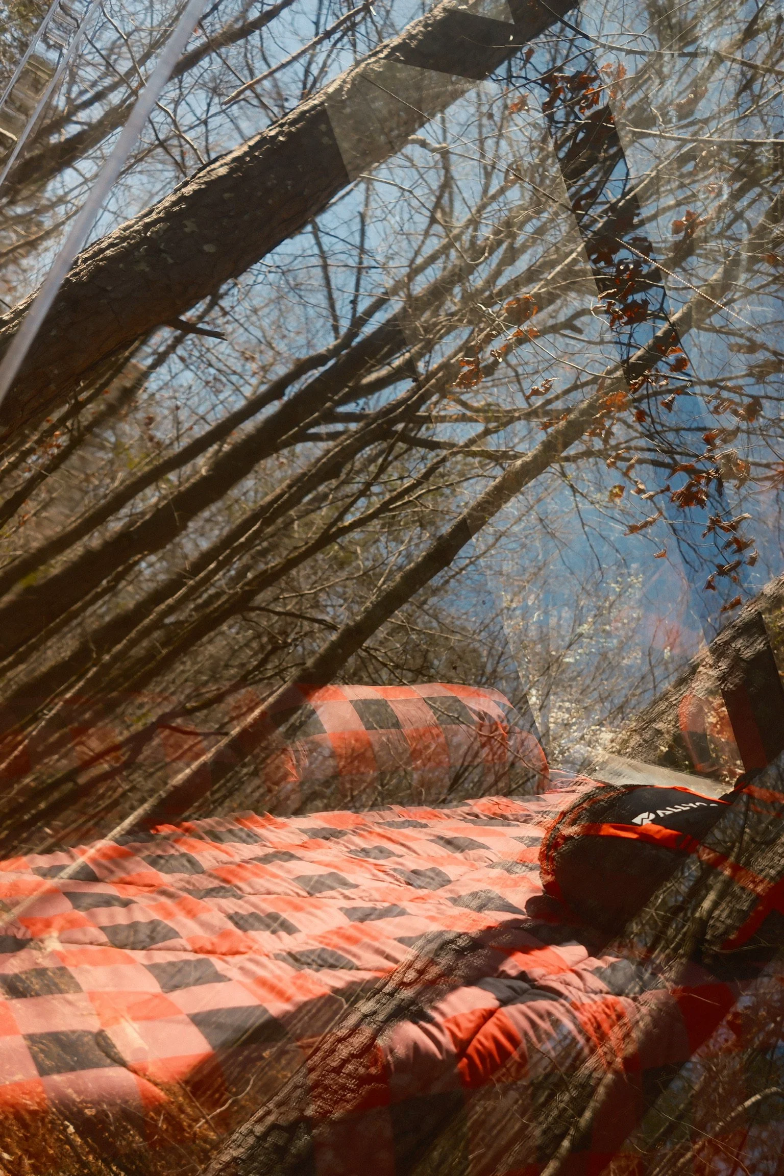 Outdoor scene with a quilted blanket and a pillow on a log, reflected on a window with trees and blue sky in the background