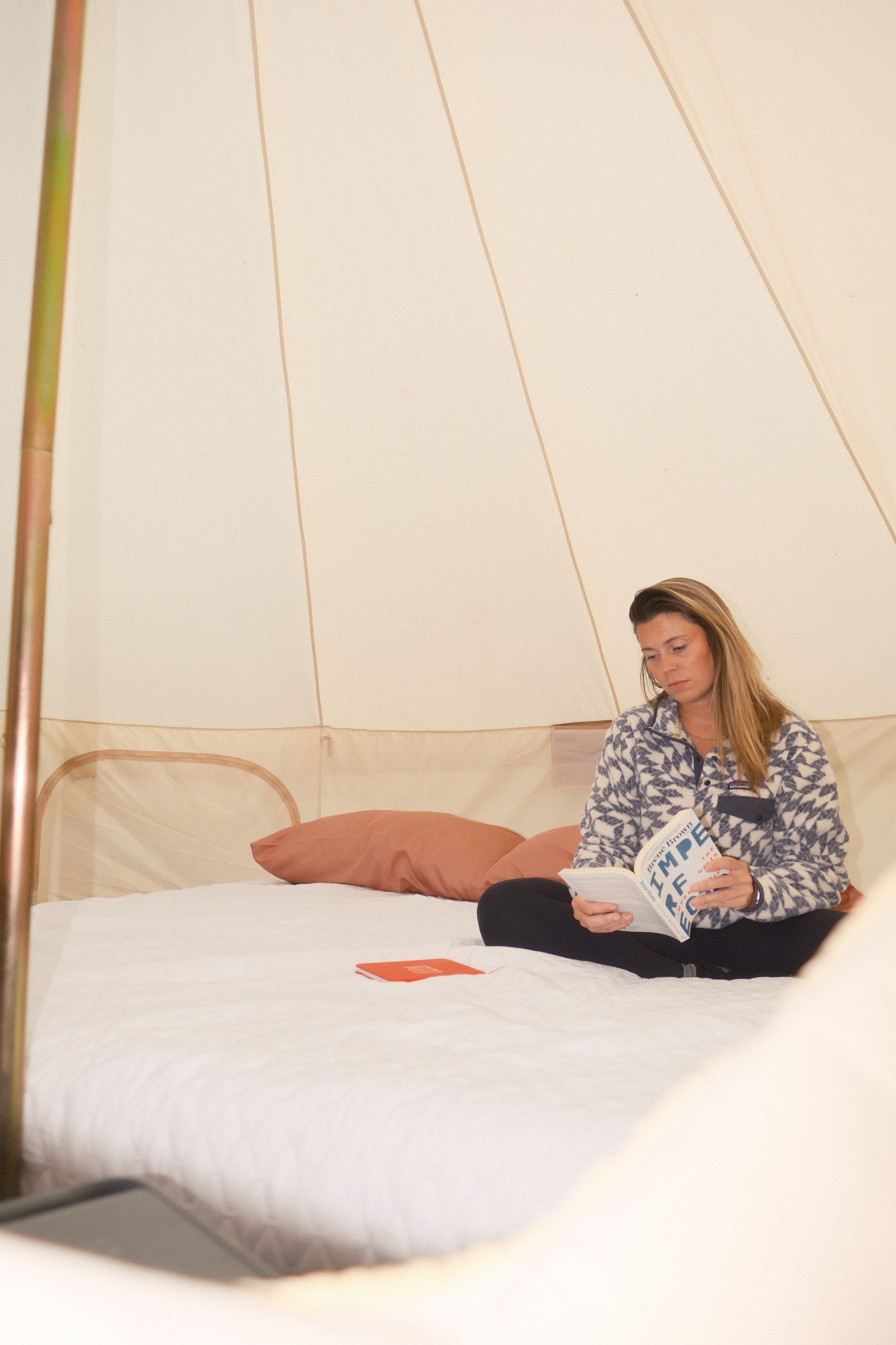 A woman sitting on a bed inside a white tent, reading a magazine. There are brown pillows and an orange book on the bed.
