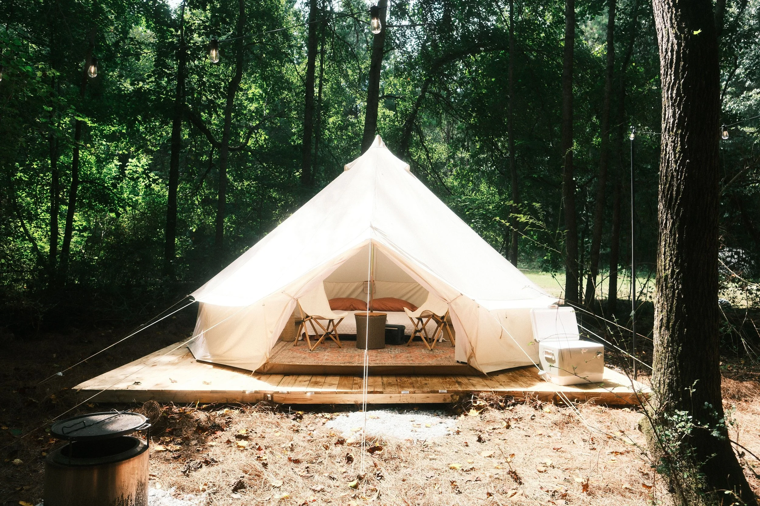 A white canvas tent set up on a wooden platform in a forest with green trees, string lights hanging above, and outdoor furniture inside.