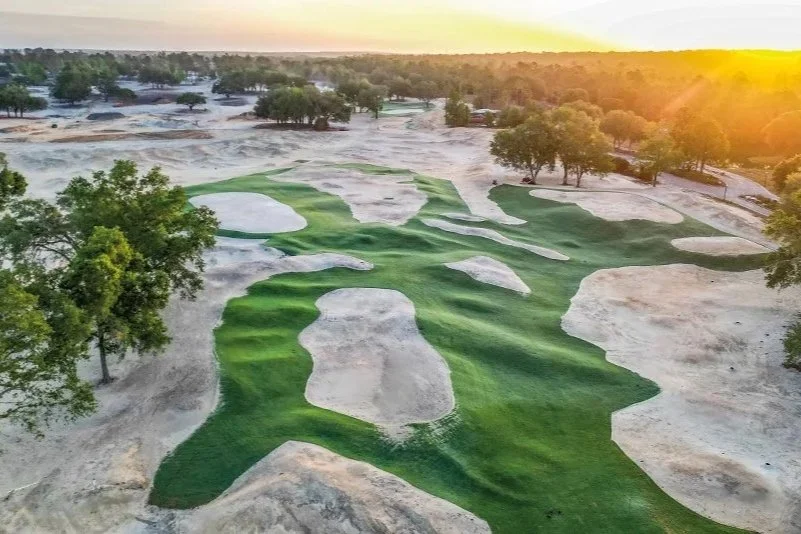 A golf course with green fairways surrounded by sandy terrain and trees, during sunset.