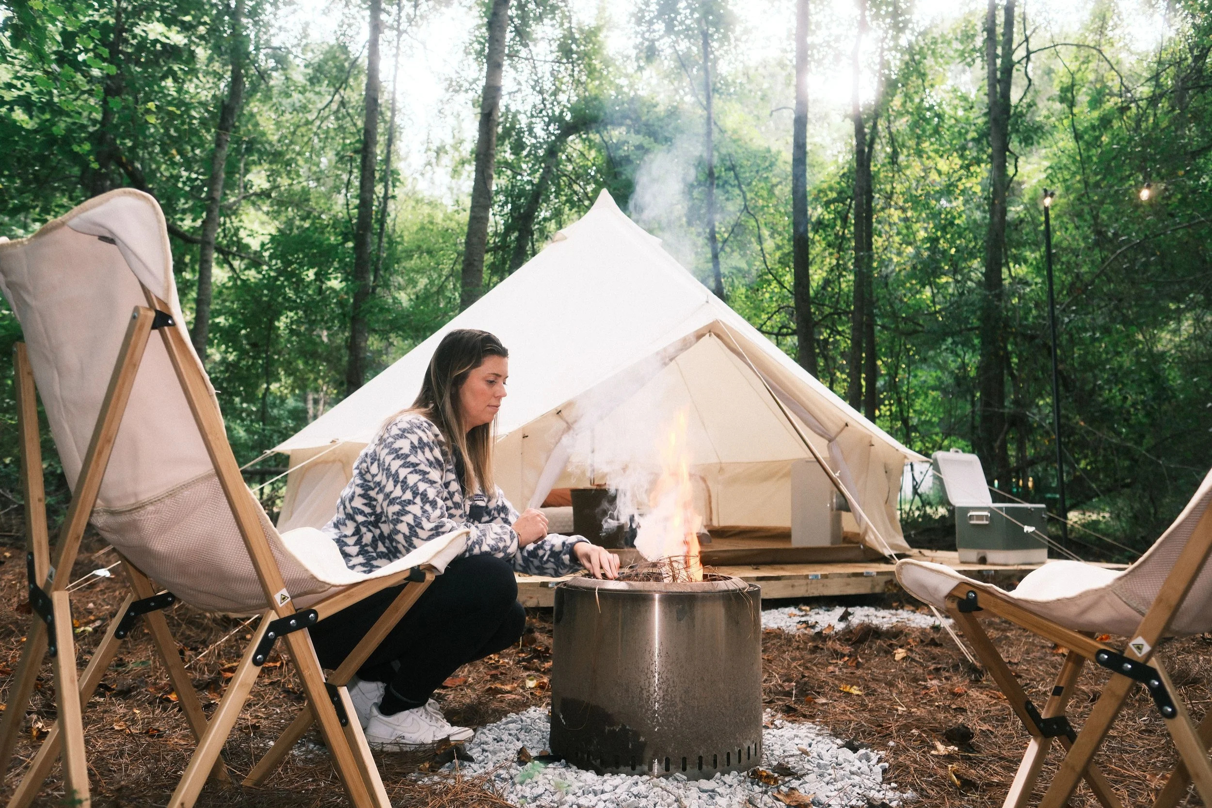 A woman sitting by a campfire in a forest, with a tent and camping chairs around her.