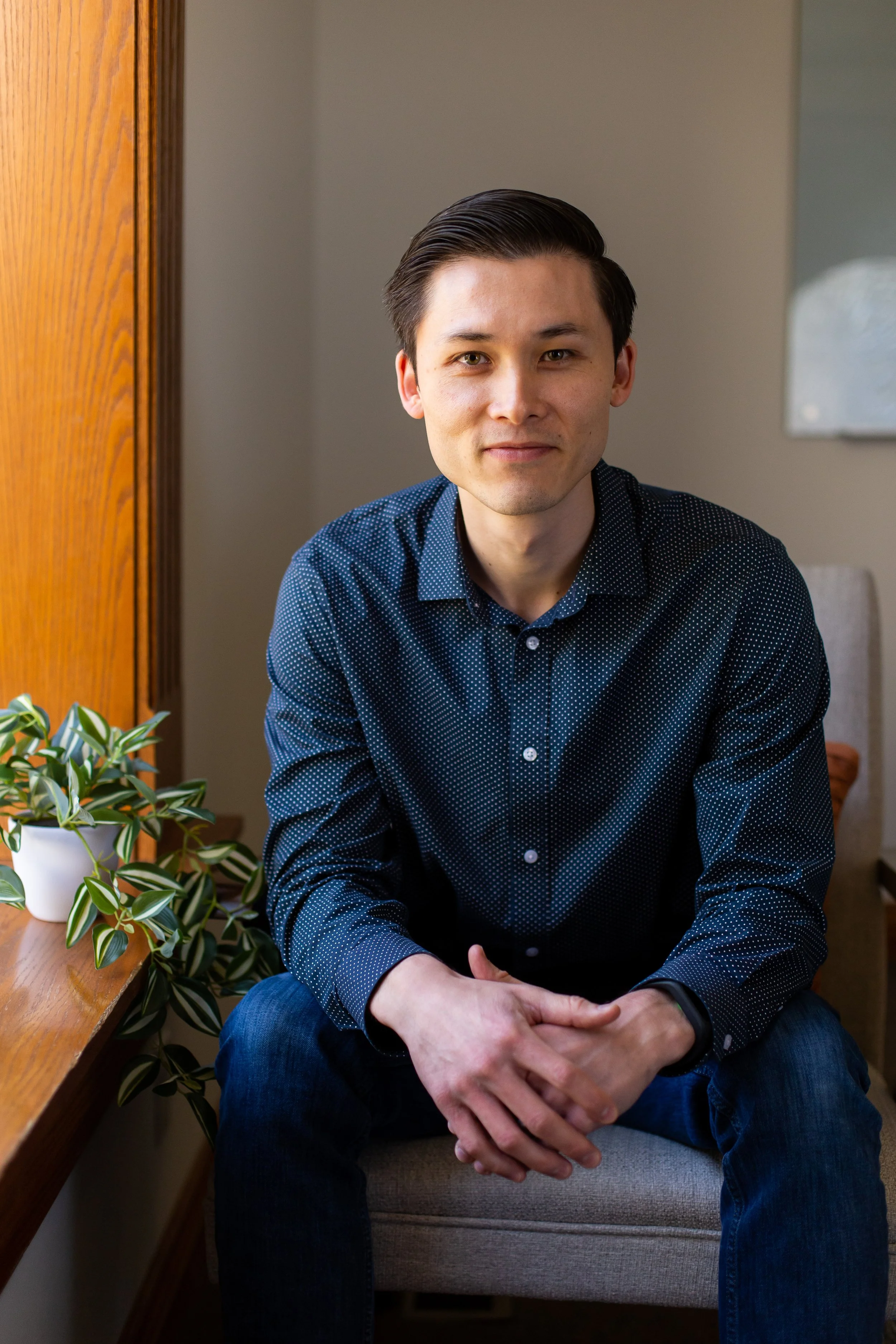 A young man with dark hair, wearing a blue patterned shirt and jeans, sitting on a beige chair in a room with a wooden door and a potted plant nearby.