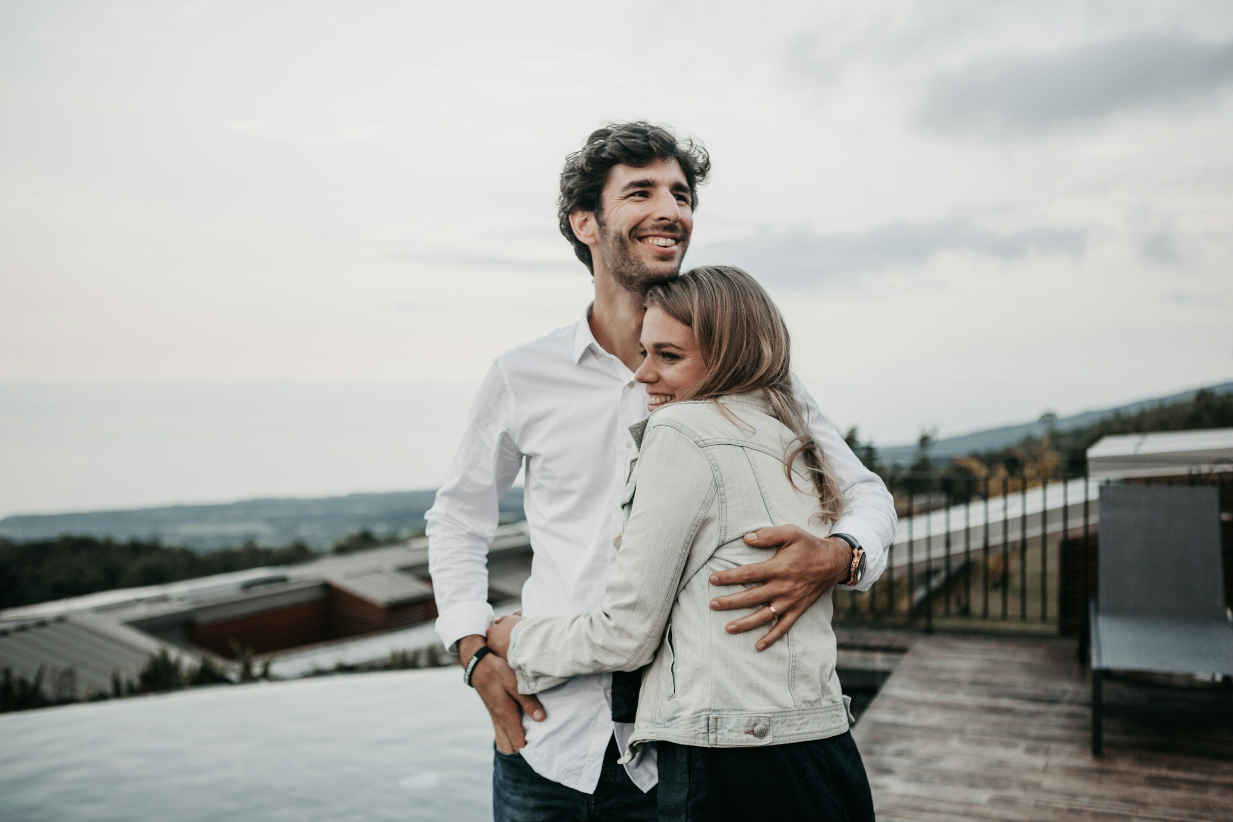 A smiling man and woman hugging outdoors on a deck with a cloudy sky and distant hills in the background.