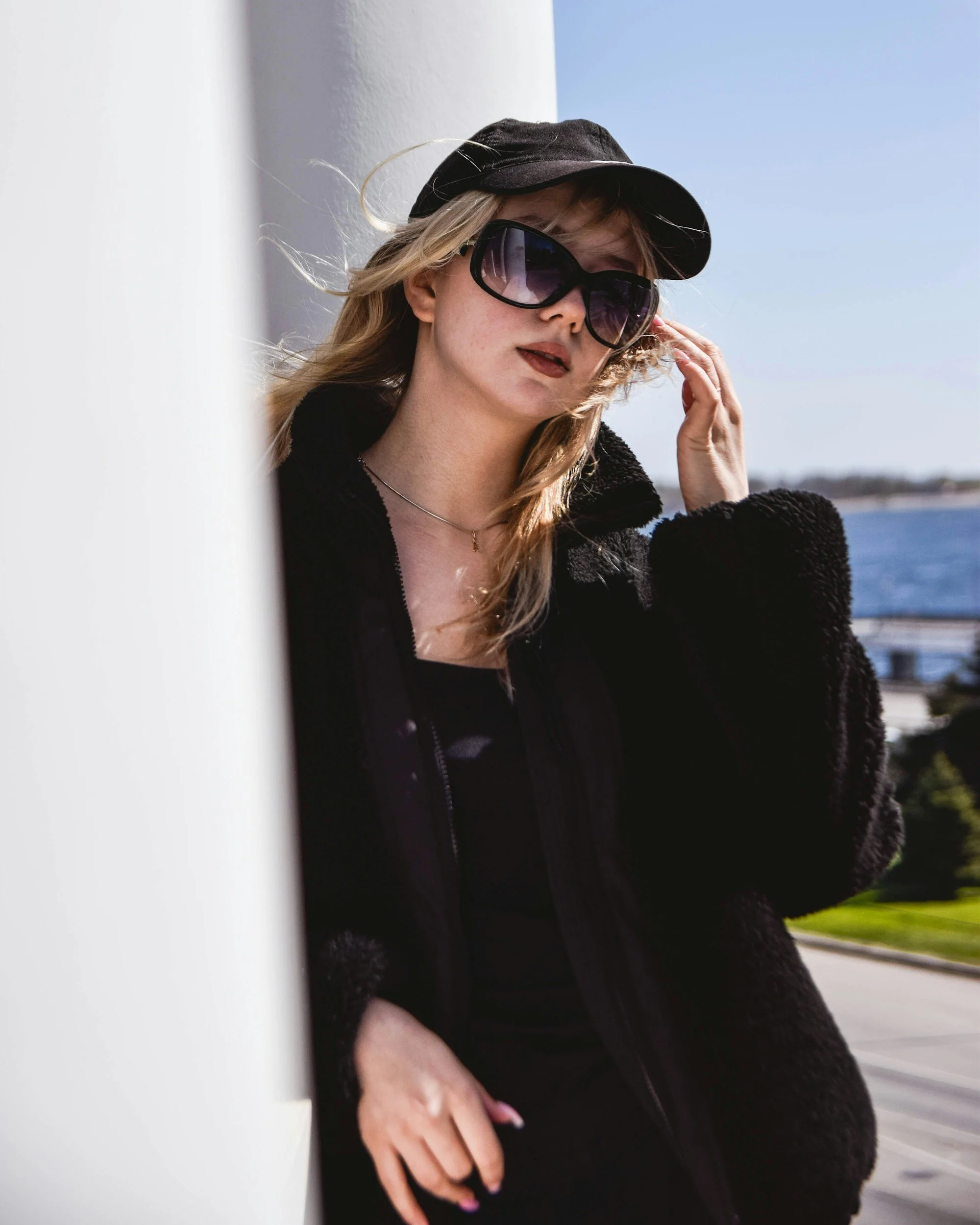 A woman with blonde hair wearing large dark sunglasses, a black cap, and a black jacket, standing outdoors near water.