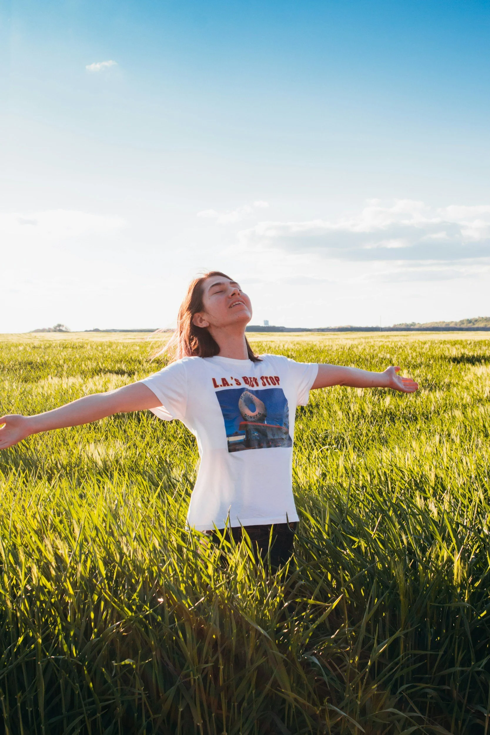 Woman standing in a grassy field with her arms outstretched and face turned upward, enjoying the sunlight under a partly cloudy sky.