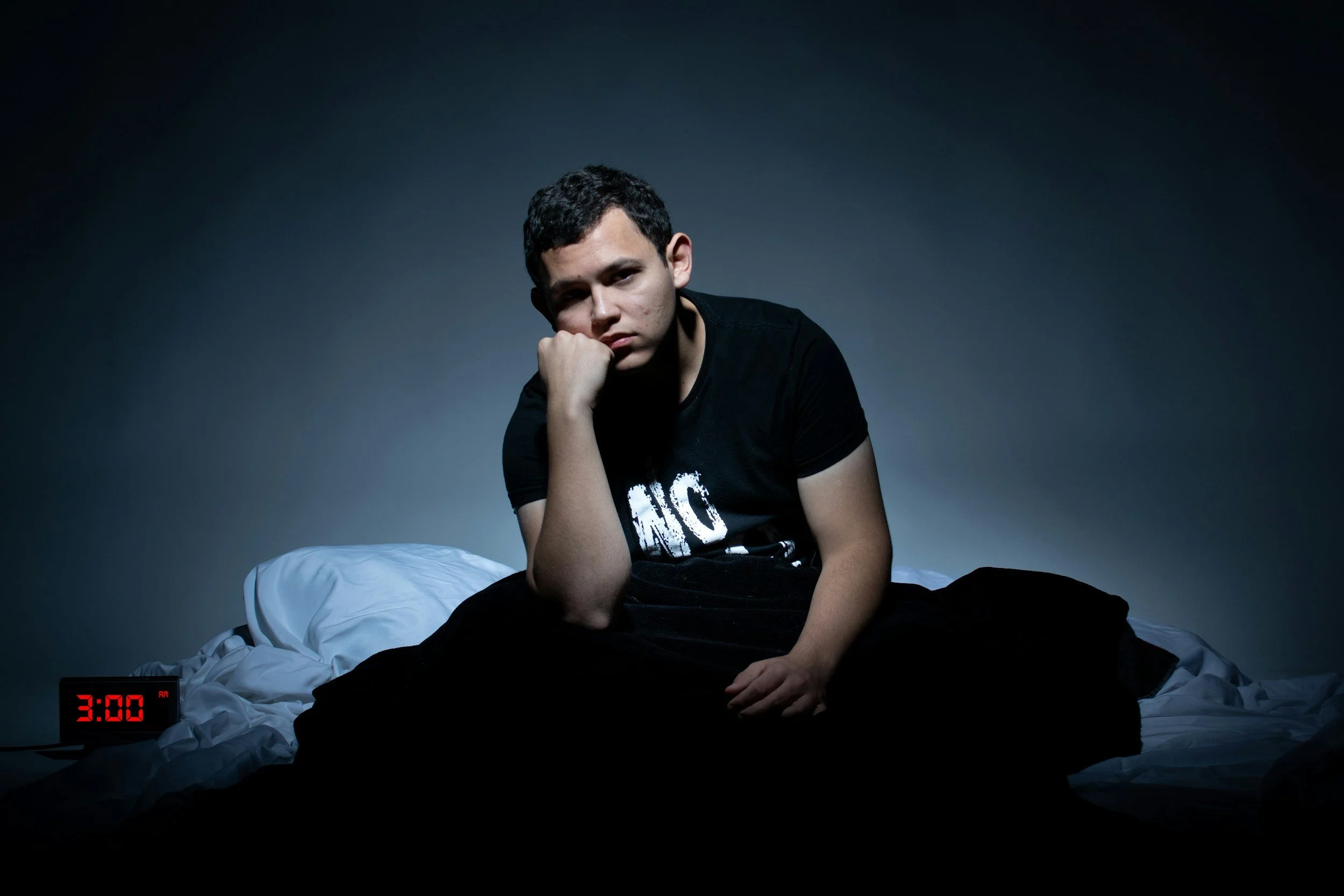 A young man sitting on a bed in a dark room with a somber expression, resting his chin on his hand, wearing a black t-shirt, and a digital clock showing 3:00 is on the bedside table.