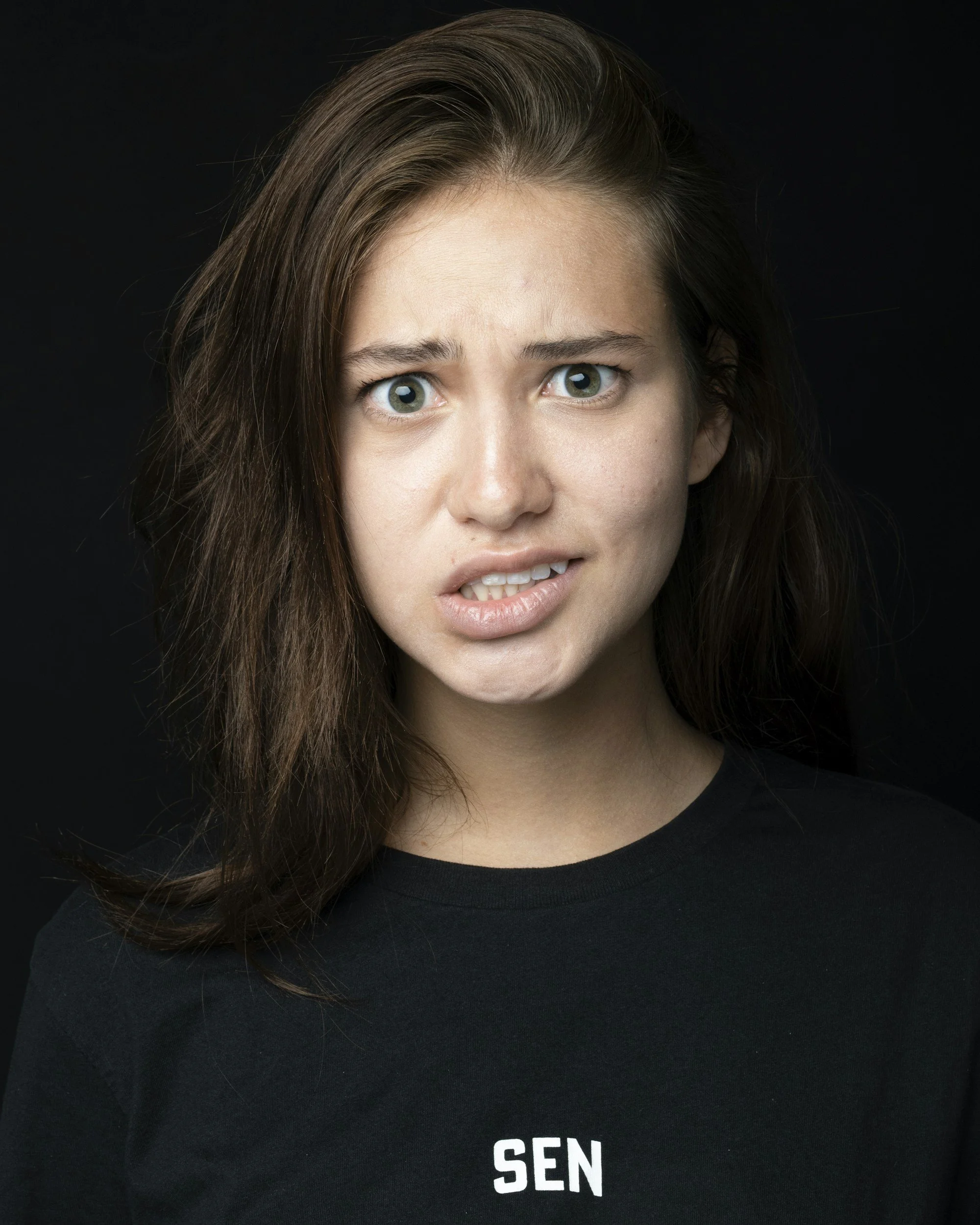 A woman with brown hair and blue eyes has a confused or puzzled expression, wearing a black shirt with partially visible white text 'SEN' on a black background.