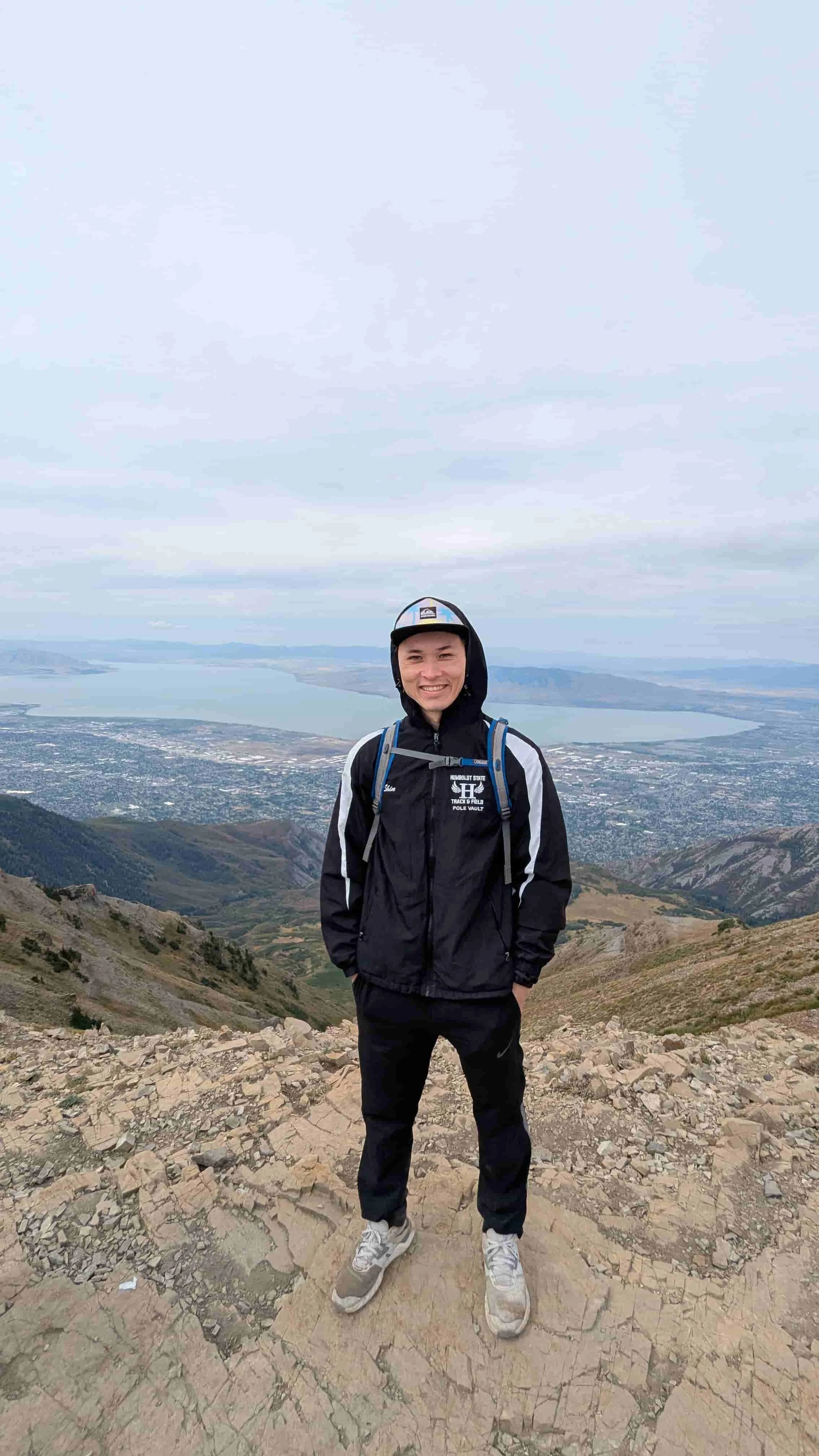 A man standing on a rocky mountain trail with a scenic view of a body of water and distant hills in the background, dressed in a black jacket, black pants, white sneakers, and a cap, smiling at the camera.