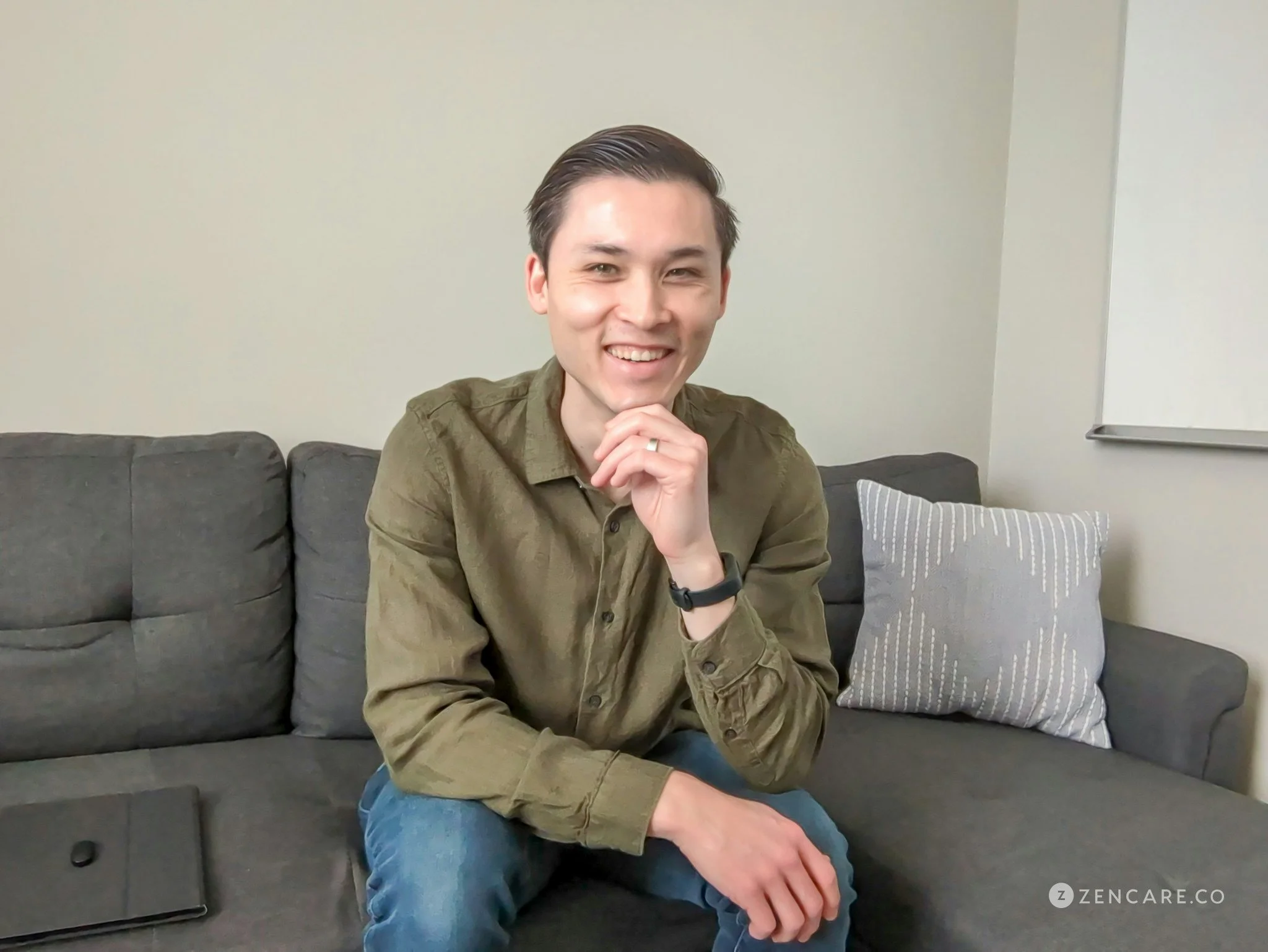 Young man with dark hair, smiling, wearing a green shirt and blue jeans, sitting on a dark gray couch in a room with beige walls, a gray and white pillow, and a closed black laptop on the couch.