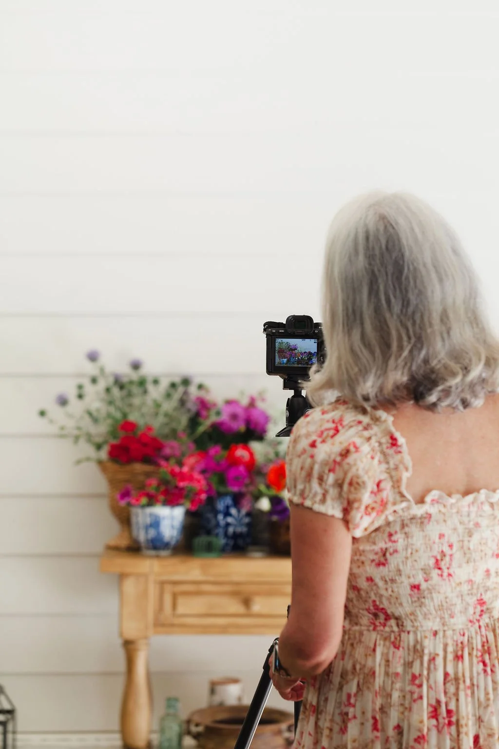 Artist Karey Bancroft taking a photo of a colorful flower arrangement on a wooden table using a camera on a tripod. (photo by Harmonie House Images)