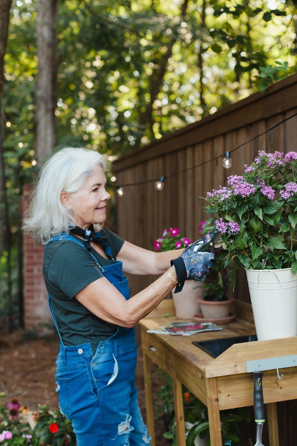 Artist Karey Bancroft working in her garden, trimming pink flowers in a white pot, wearing gloves, a black shirt, and jeans, standing beside a wooden gardening station with various plants and gardening tools. (photo by Harmonie House Images)