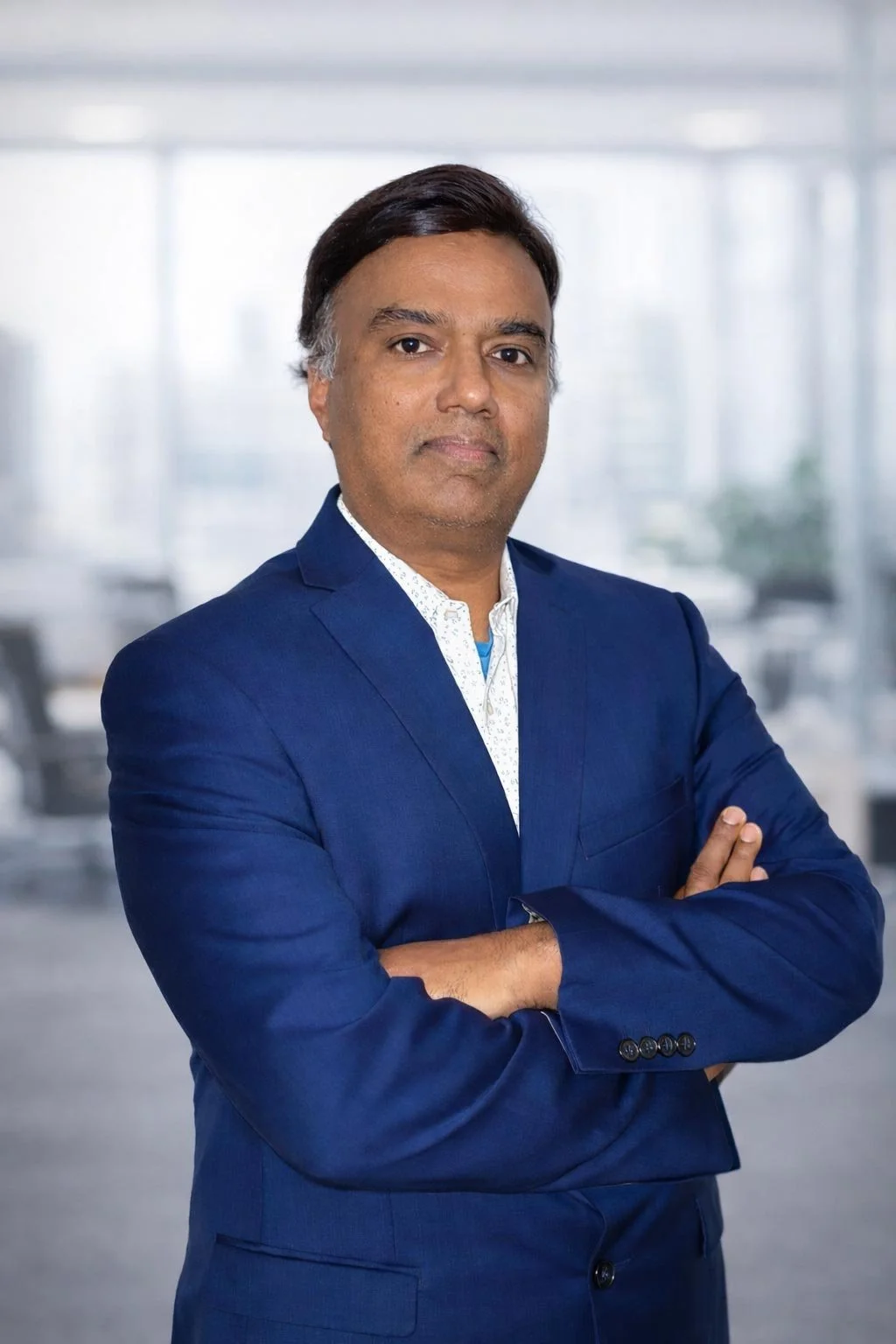 A professional man in a blue suit with arms crossed, standing in a bright modern office with blurred background.