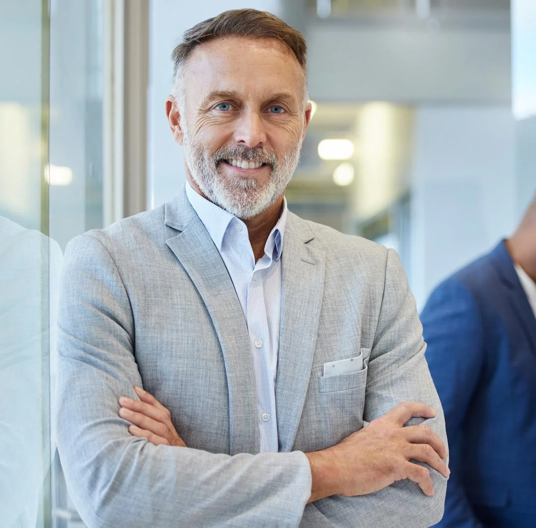 A middle-aged man with a beard and short hair, wearing a light gray blazer and a white shirt, smiling confidently with arms crossed, standing in a modern office setting.