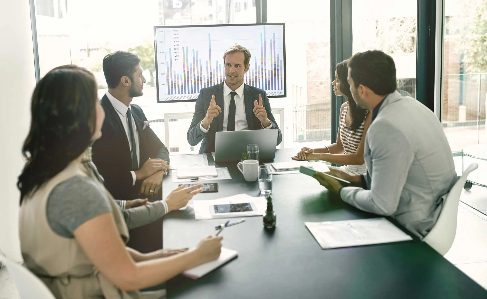 A man in a suit is giving a presentation to six colleagues seated around a conference table, with a large chart displayed on a screen behind him.