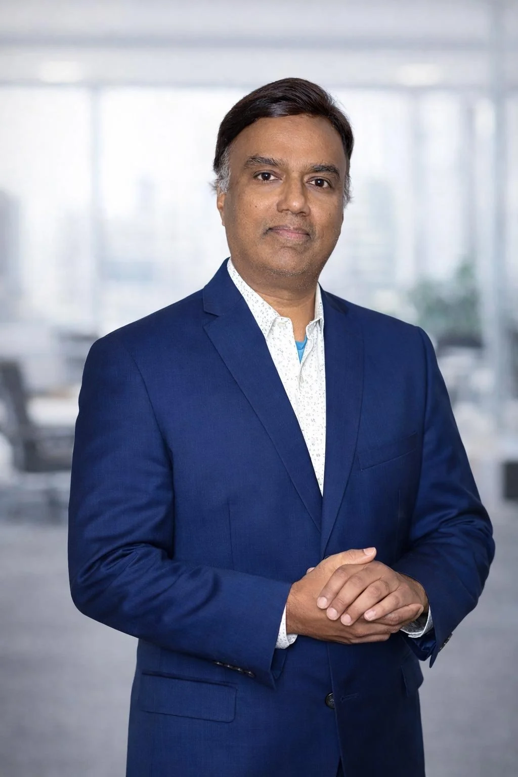 A professional man in a blue suit standing with his hands clasped in front of him in an office environment.