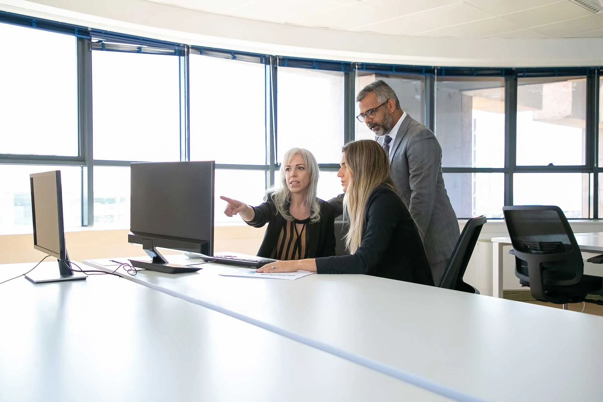 Three professionals discussing work at a modern office with large windows and two computer monitors.