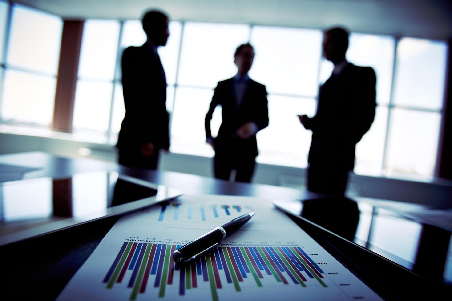 Silhouettes of three business professionals in a meeting room with large windows, with a table inset showing printed charts and a pen in the foreground.