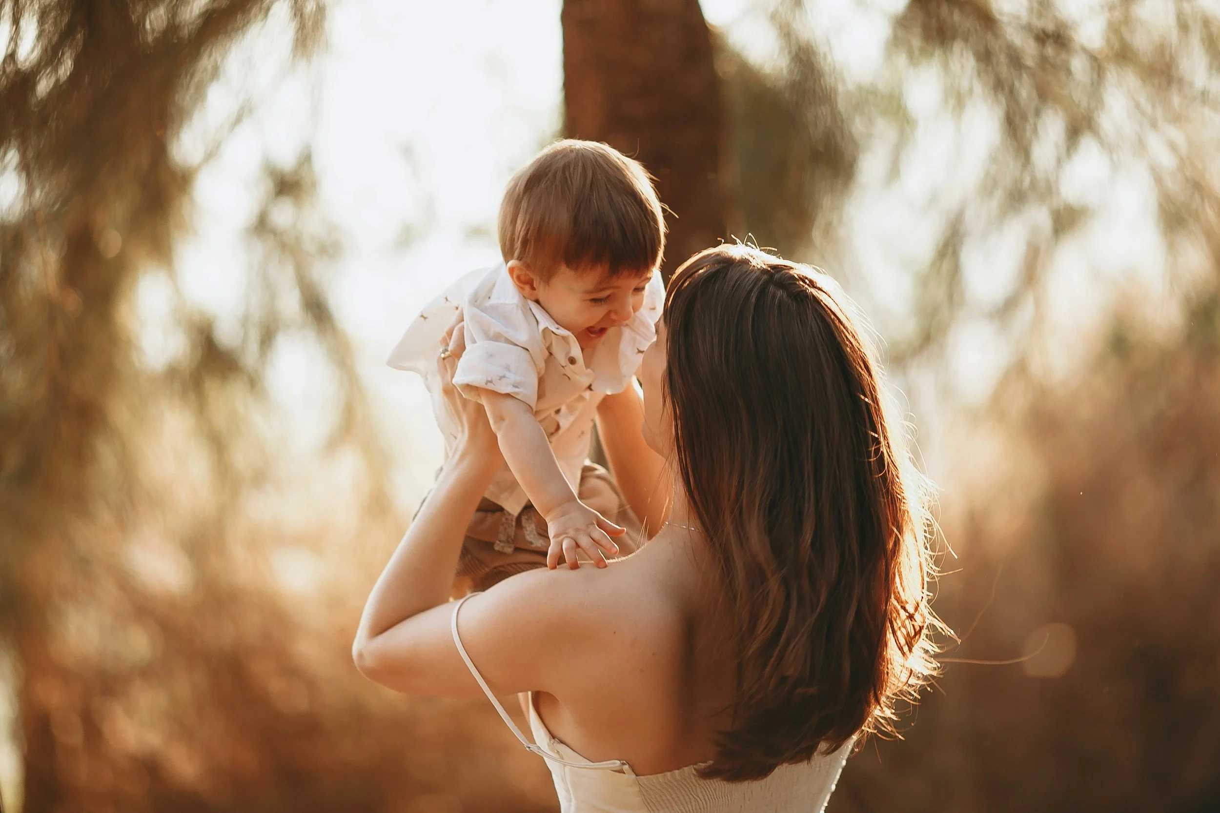A woman lifting a young child in the air outdoors during golden hour with trees in the background