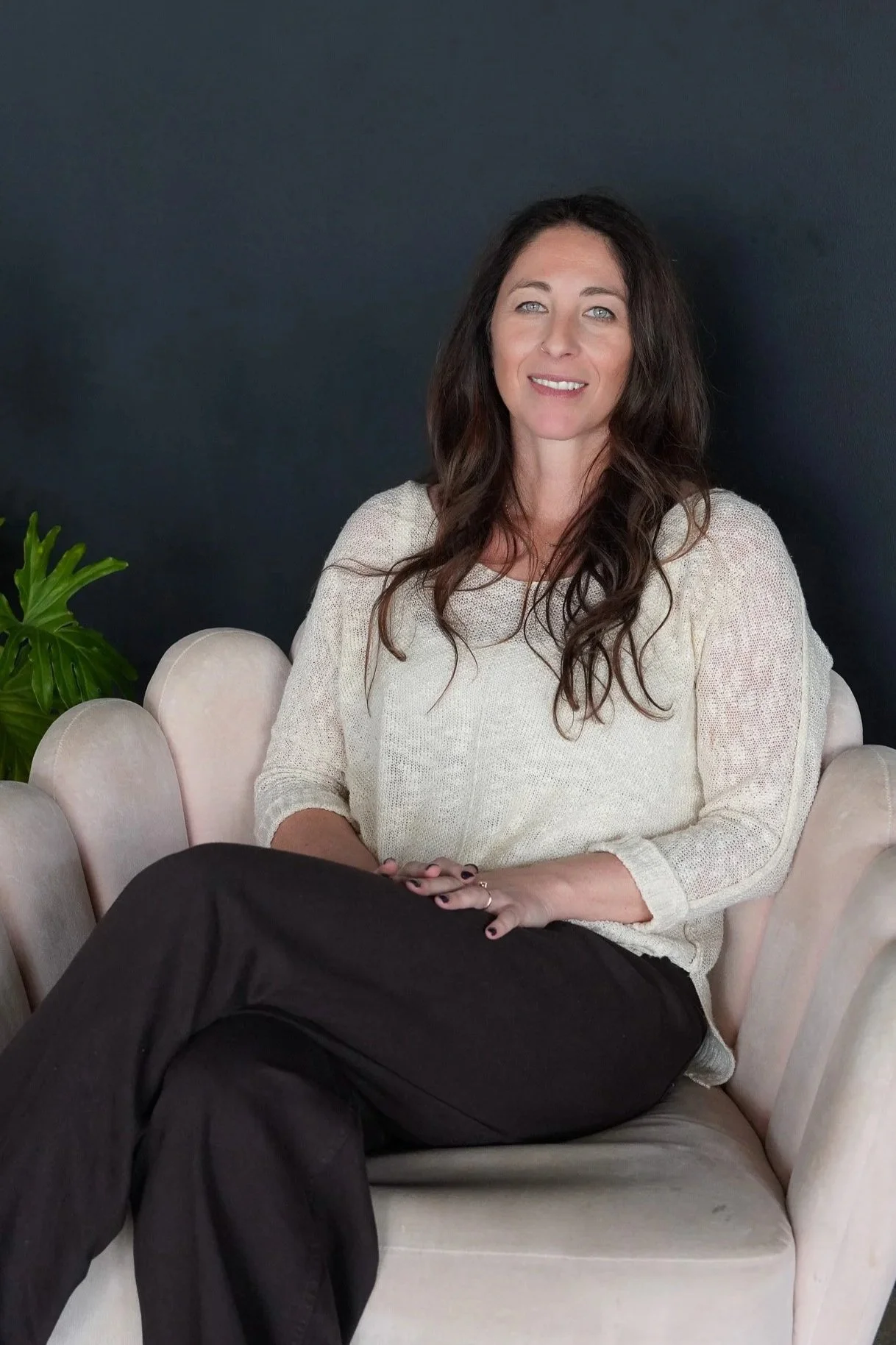 A woman with long dark wavy hair and blue eyes sitting on a light-colored velvet chair, smiling at the camera, wearing a cream-colored sweater and black pants, with a dark wall and a green plant in the background.