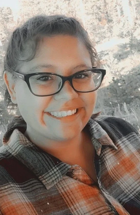 A woman smiling, wearing glasses and a plaid shirt, outdoors with trees in the background.