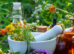 Garden scene with a mortar and pestle, fresh herbs, flowers, glass bottles, and a dropper, set outdoors surrounded by greenery and plants.