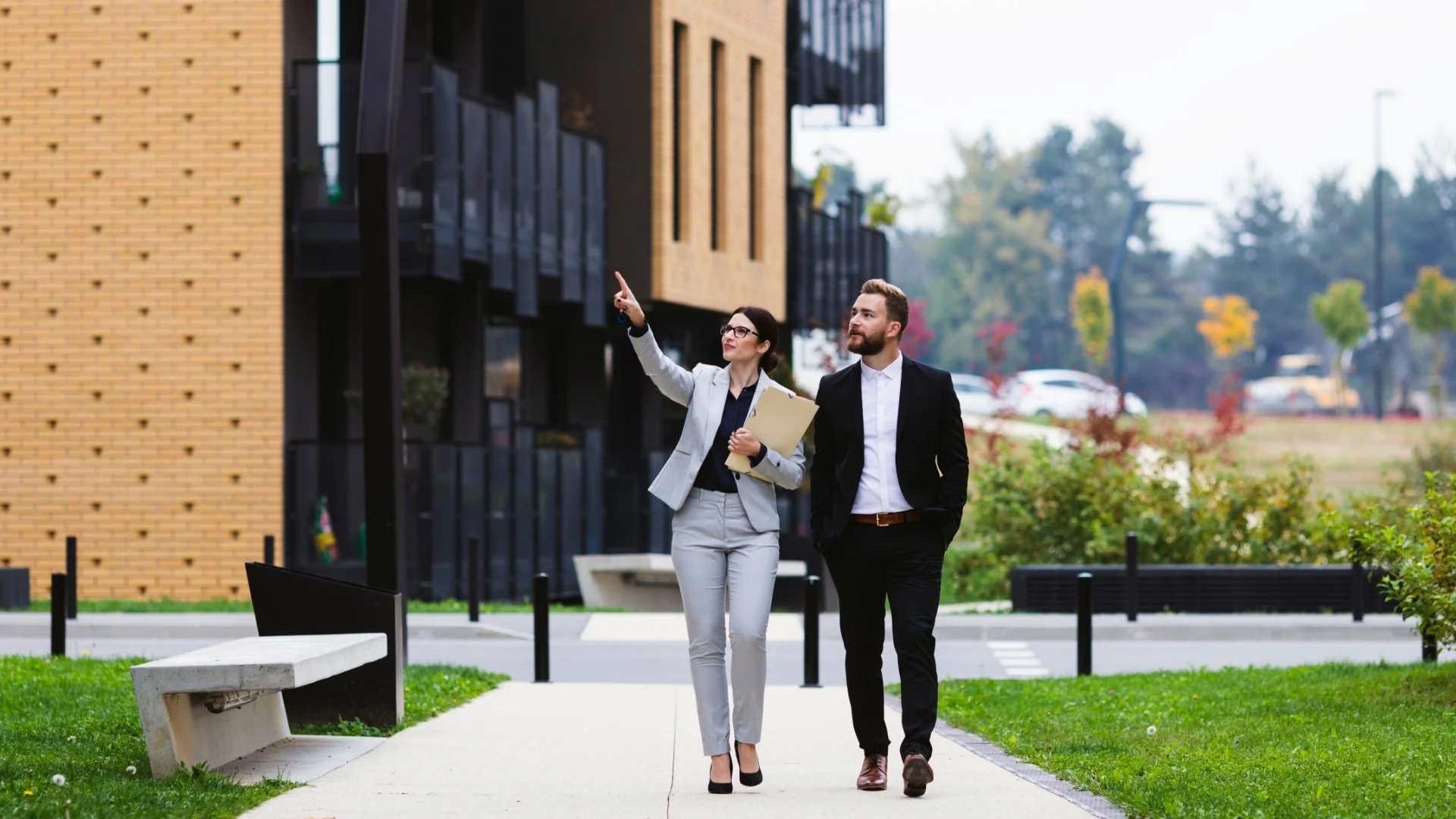 Free Home Value Now - Two business professionals, a woman and a man, walking on a sidewalk outside a modern building. The woman is pointing ahead and holding a folder, while the man listens. The background features trees, cars, and a landscaped area.