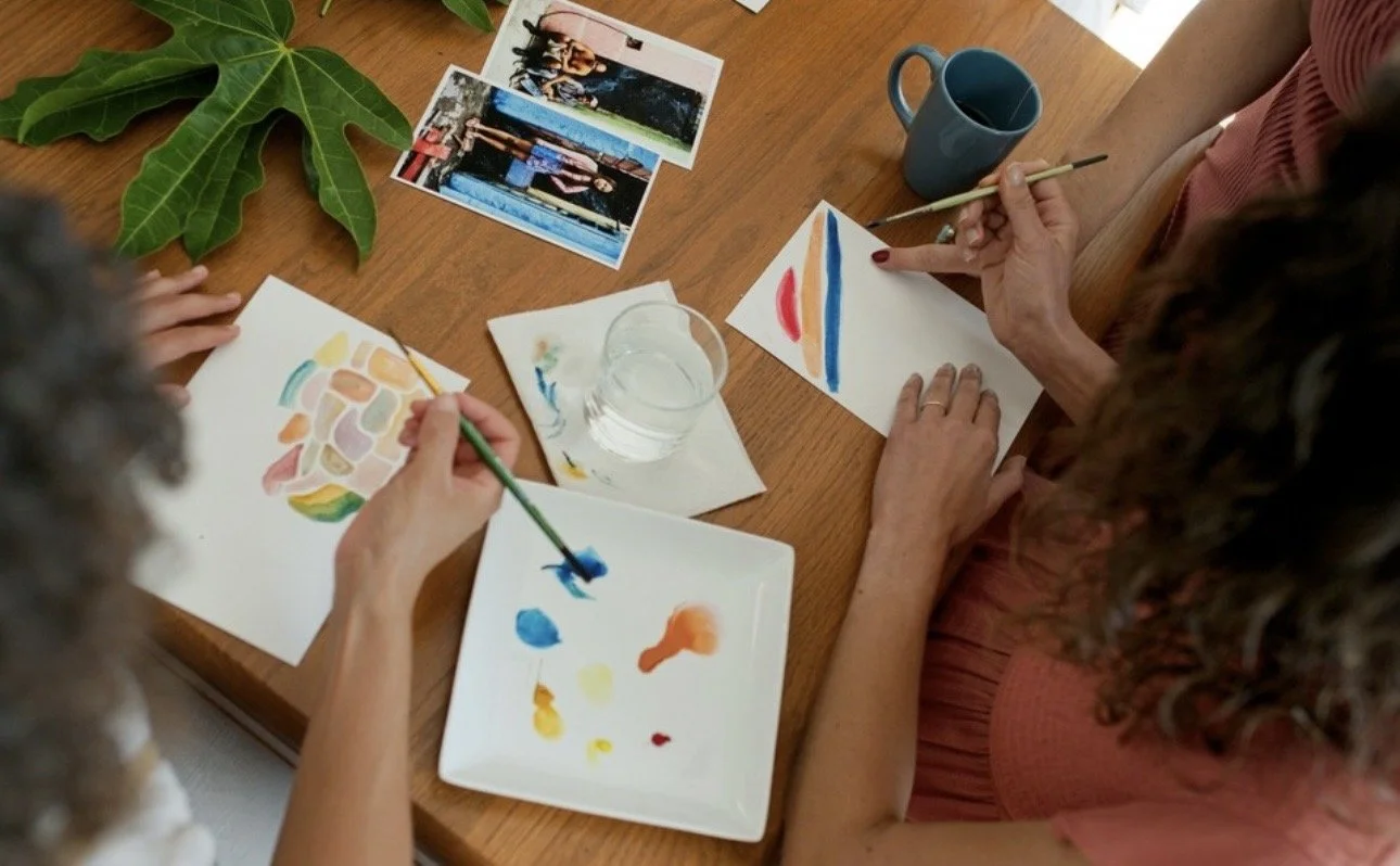 Two women painting watercolor pictures at a wooden table, with one woman painting an abstract rainbow design and the other painting colorful shapes, surrounded by photographs, a large green plant leaf, and mugs.