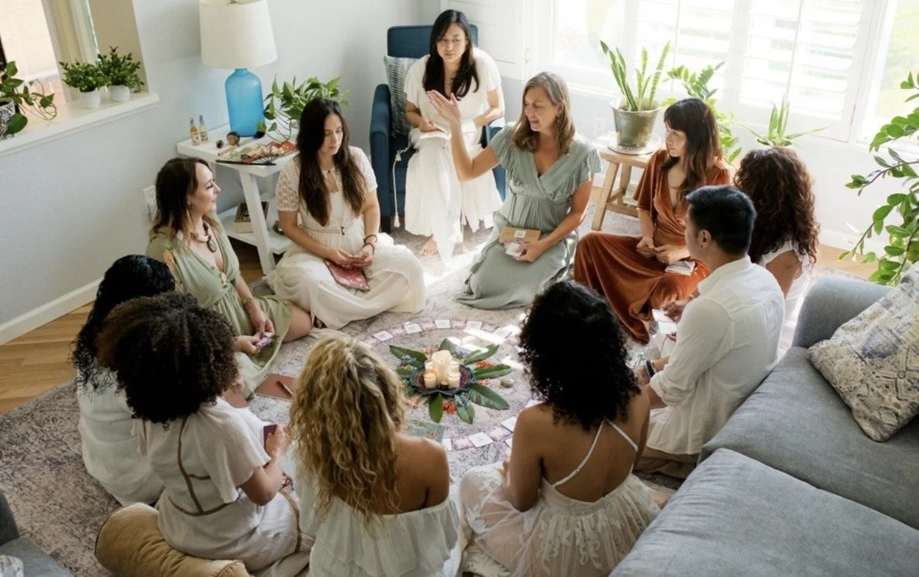 A group of women and a man sitting on a circular arrangement of candles and flowers in a living room, participating in a spiritual or meditation session.