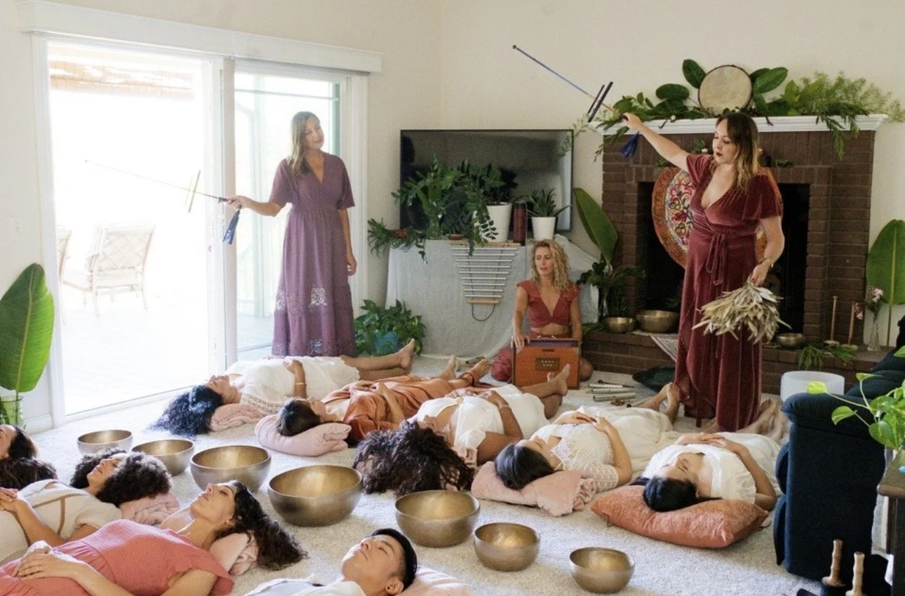 Women lying on the floor with participating in a sound bath with Tibetan bowls, sound wands, and a shurti box. Three women in dresses faciltate the experience in a cozy living room with plants and a fireplace.