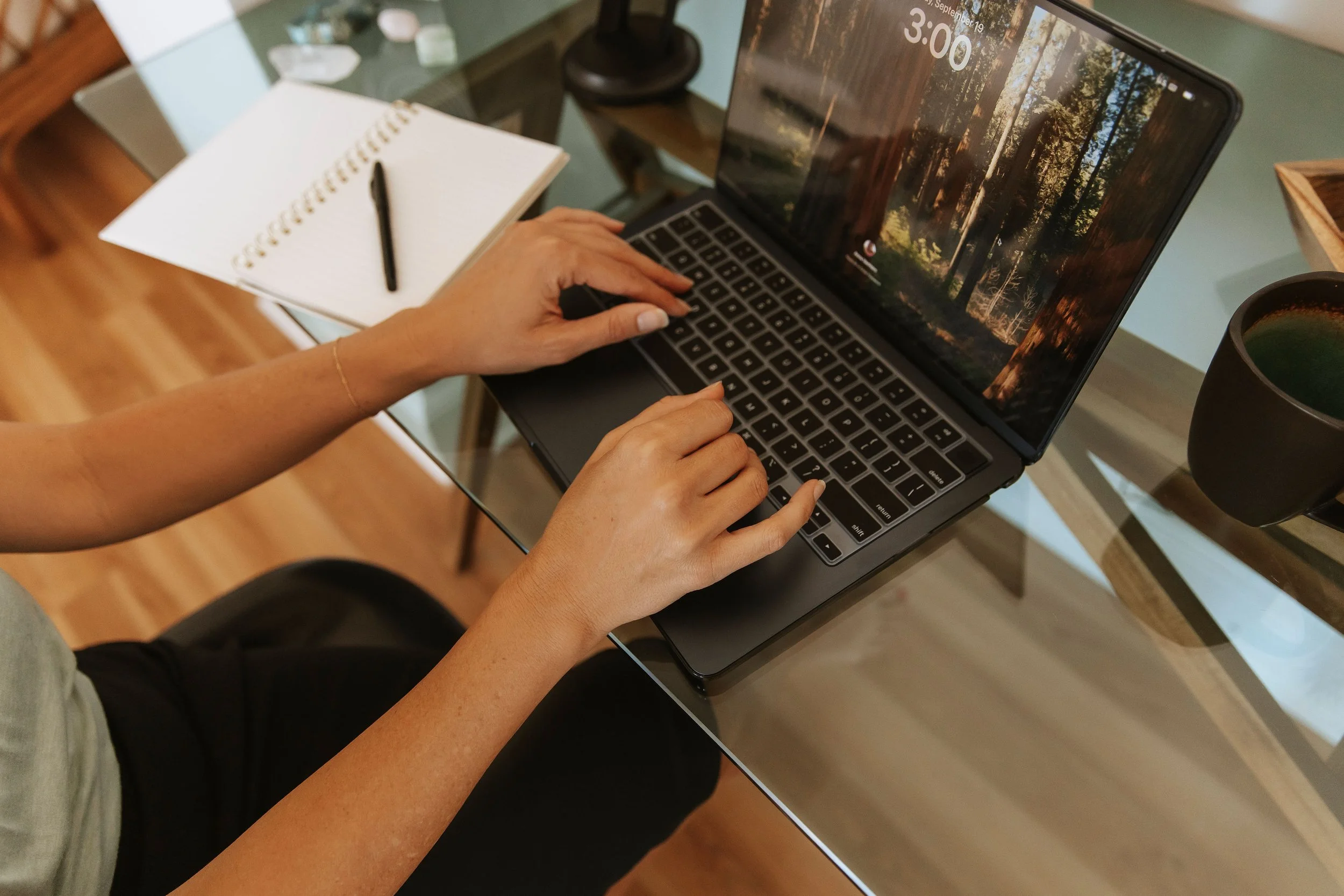 A person with light skin and a watch on their wrist is sitting at a glass table, typing on a laptop with a forest scene on the screen. There is a notebook with a pen on it, a black mug, and some decorative items in the background.