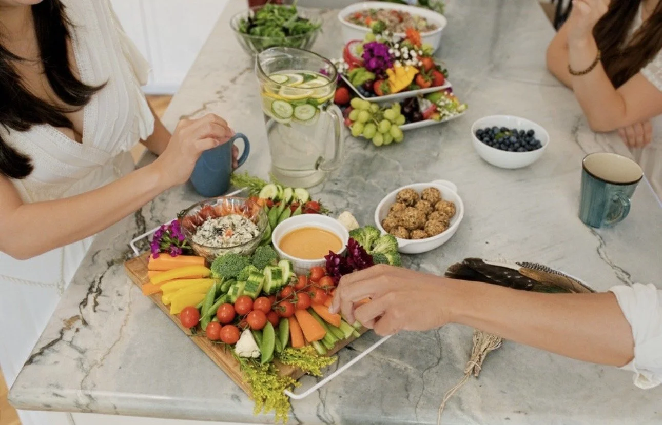 People having a party with a large marble countertop filled with fresh vegetables, fruit platter, bowls of blueberries, mixed greens, and other dishes, along with a pitcher of infused water, mugs, and small bowls of dips.