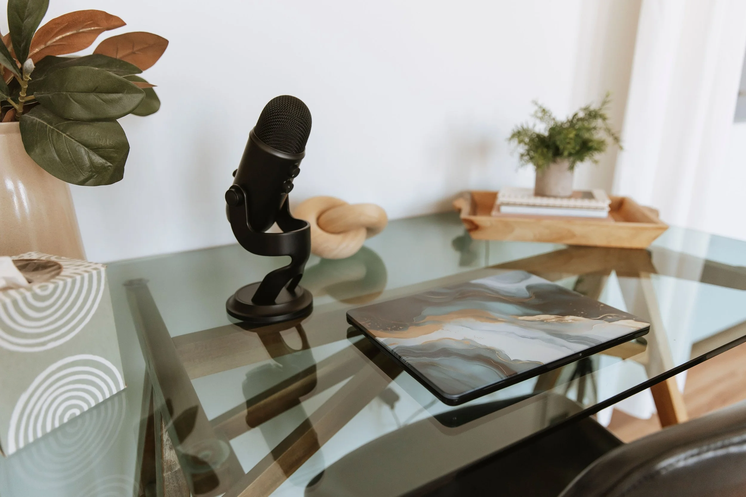 A home decor setup on a glass table with a black microphone on a stand, a beige vase with green and brown leaves, a small wooden tray with a potted plant and a stack of books, and a laptop with a marbled cover.