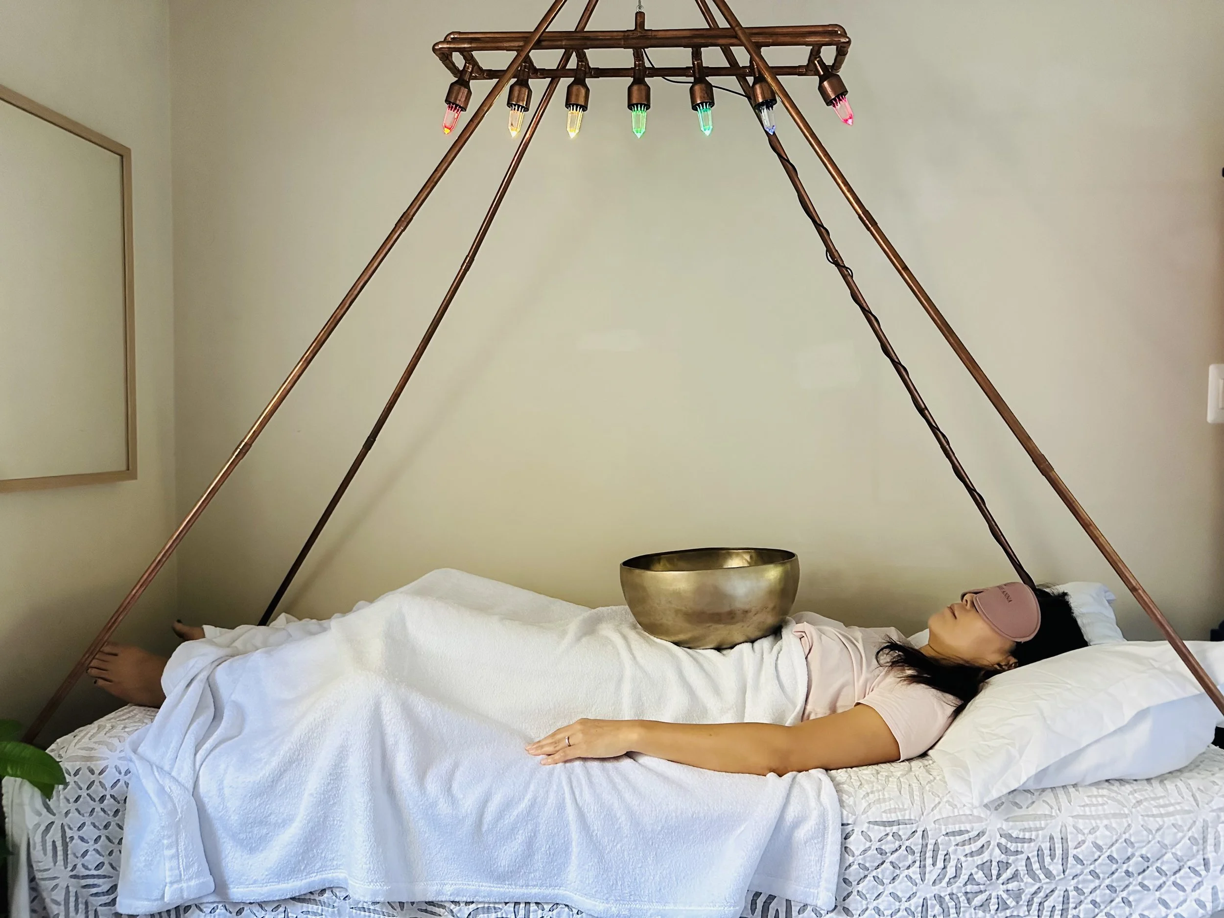 A woman lies on a vibroacoustic therapy bed with a white sheet, wearing a pink sleep mask, surrounded by a copper pyramid structure with colored Vogel crystal lights, and a Tibetan singing bowl on her stomach.