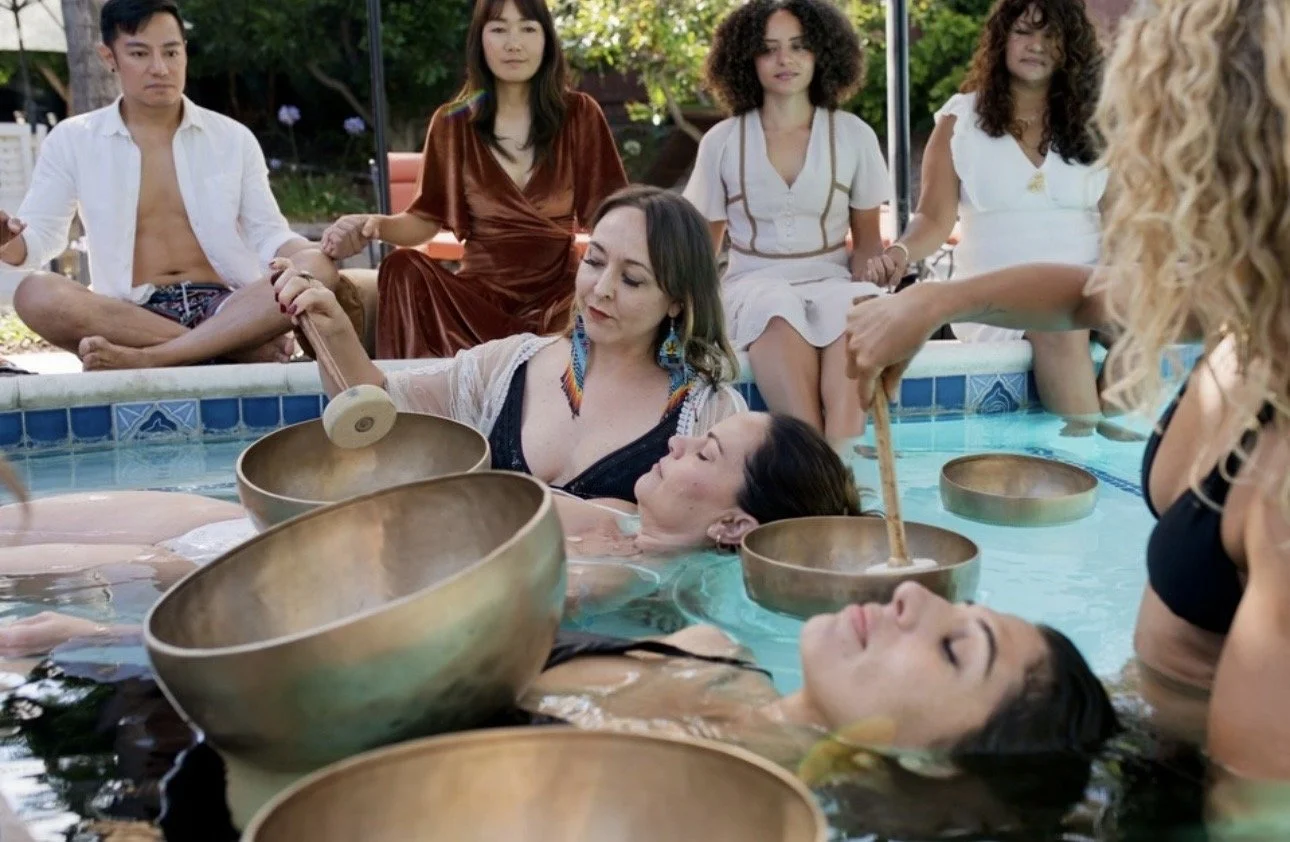 People participating in a sound bath meditation in an outdoor pool, with several people sitting on the edge and others lying in the water, holding singing bowls and feeling the healing vibrations.